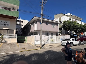 Un edificio de varios pisos con una entrada protegida por una reja, una camioneta blanca estacionada cerca y un cielo azul despejado.