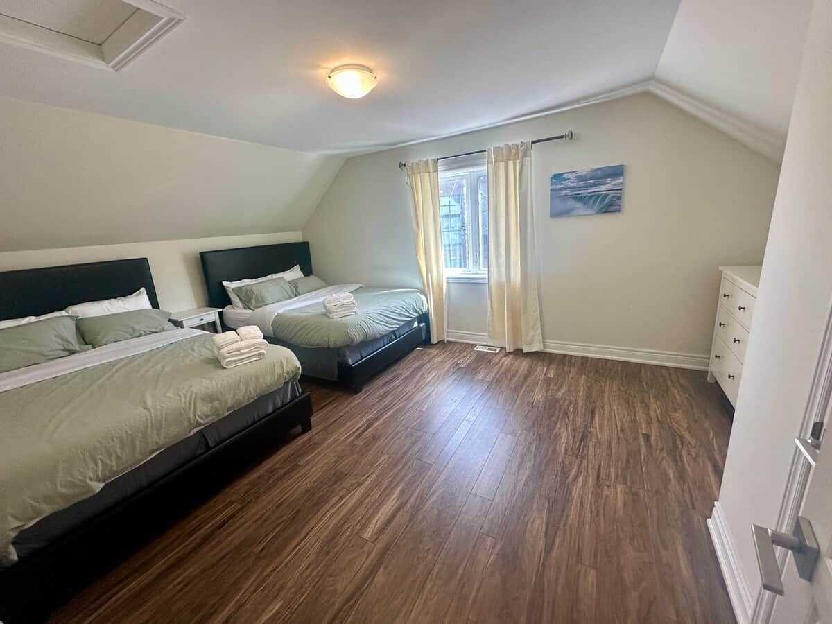 A bedroom with two beds, a window with curtains, a painting on the wall, and a wooden floor.