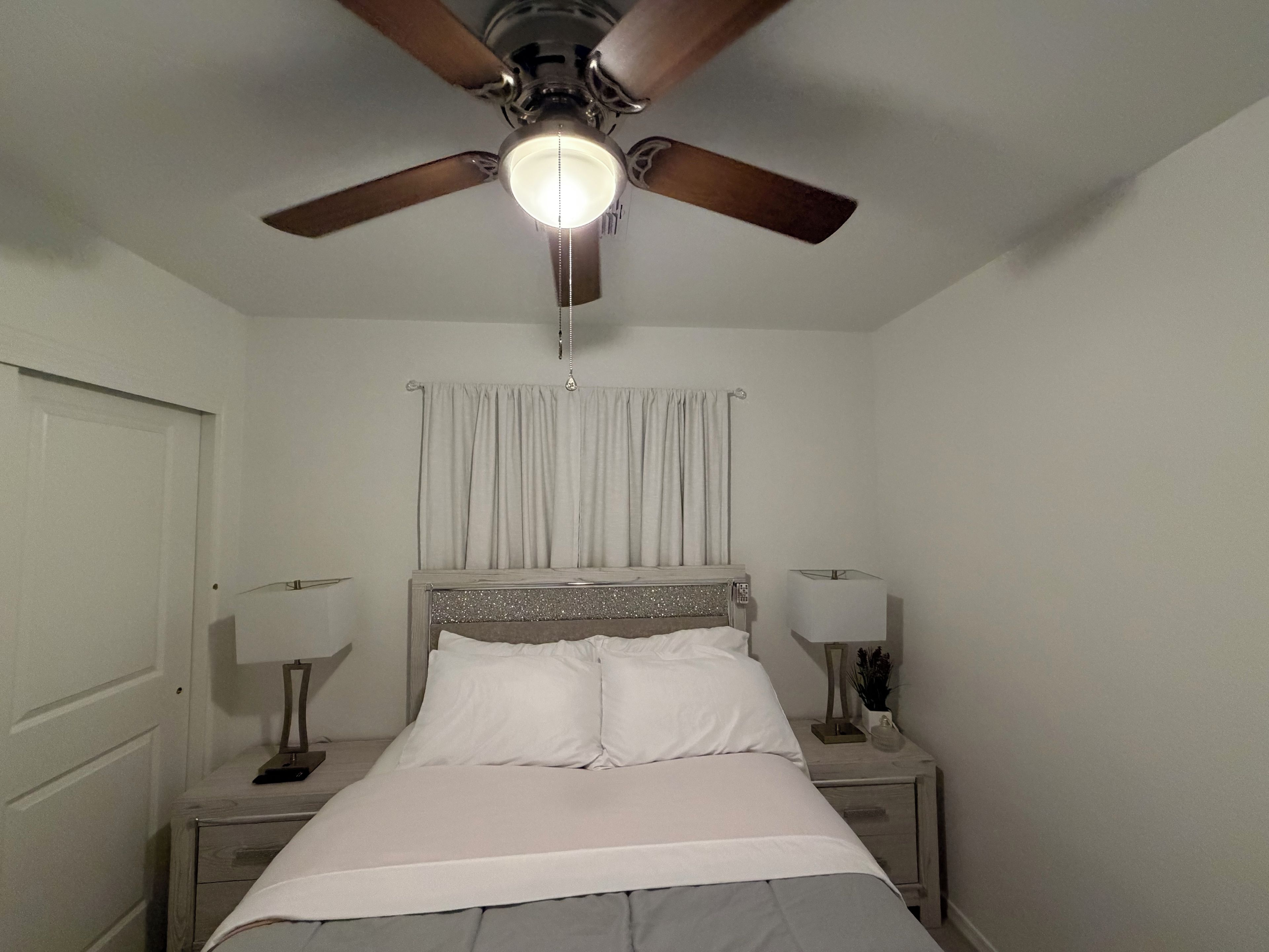 A bedroom with a bed, two bedside tables, a ceiling fan, and a window with curtains.