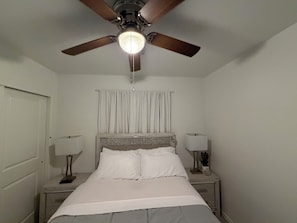 Un dormitorio con una cama, dos mesitas de noche, un ventilador de techo y una ventana con cortinas.