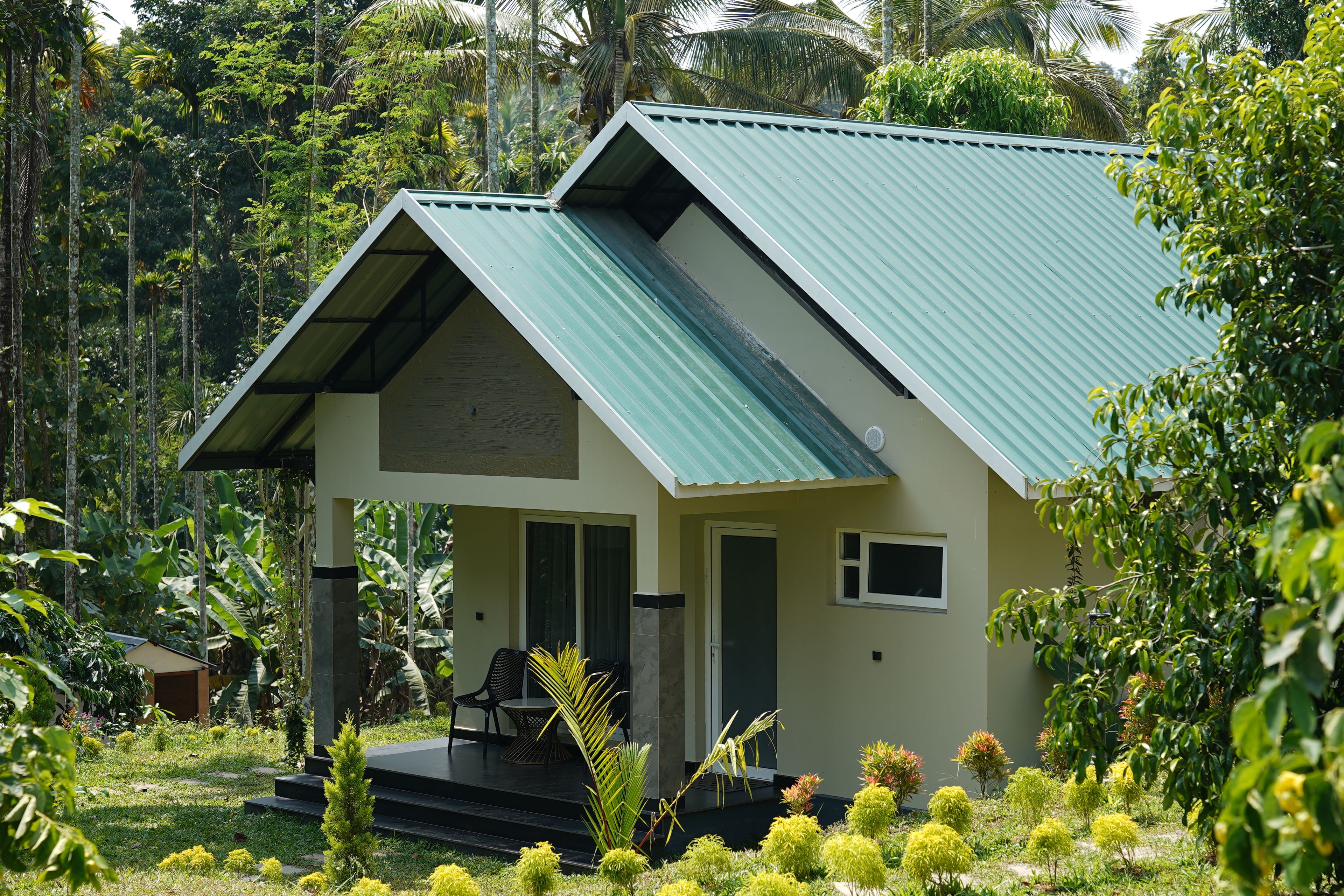 A two-story house with a green roof, a porch with chairs, and a garden with various plants.