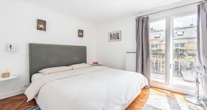 Un dormitorio con una cama grande, una mesita de noche, una ventana con cortinas y vistas a edificios.