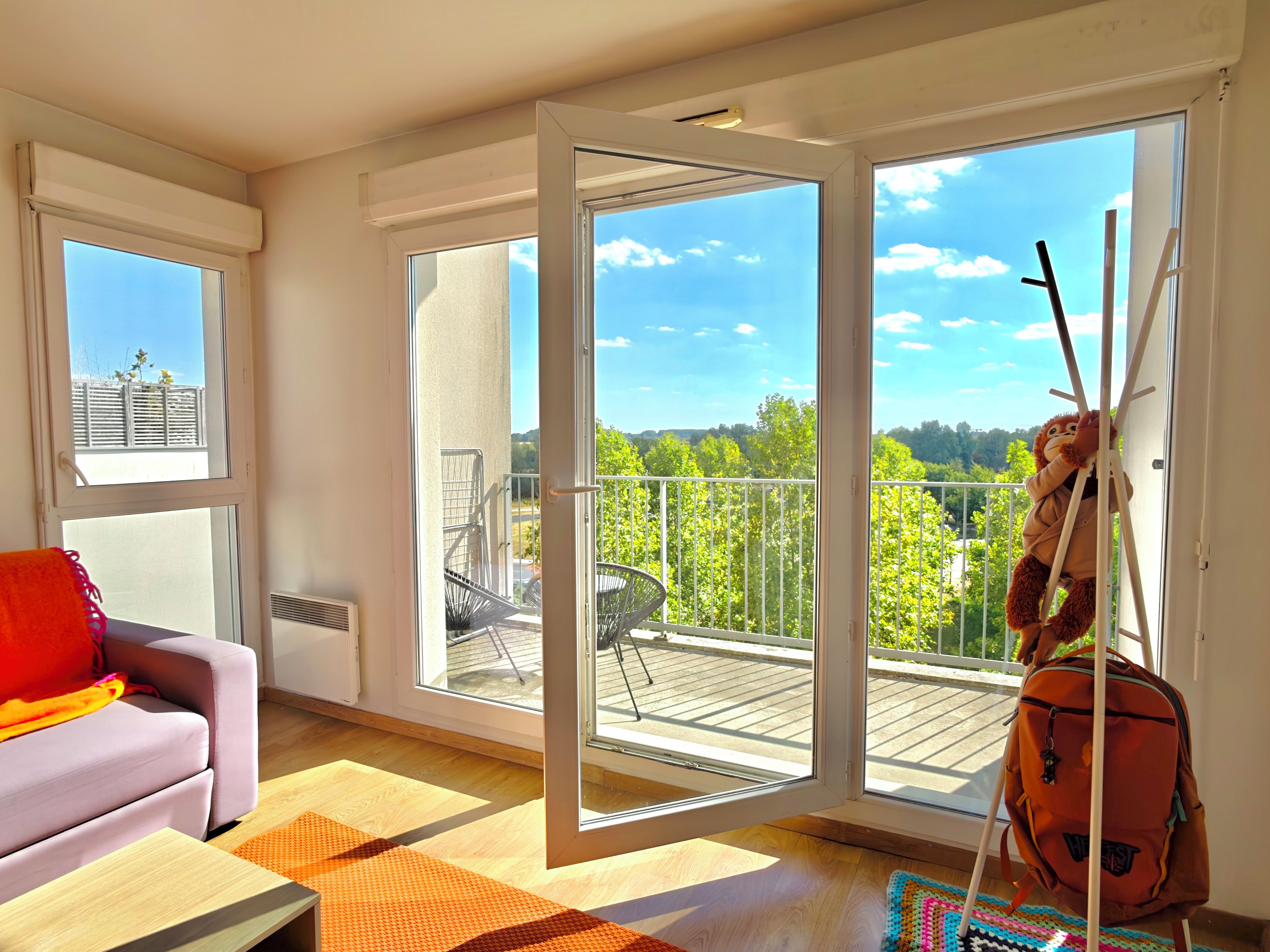 A modern living room with a sofa, a colorful rug, and a large window with a view of a balcony and greenery.