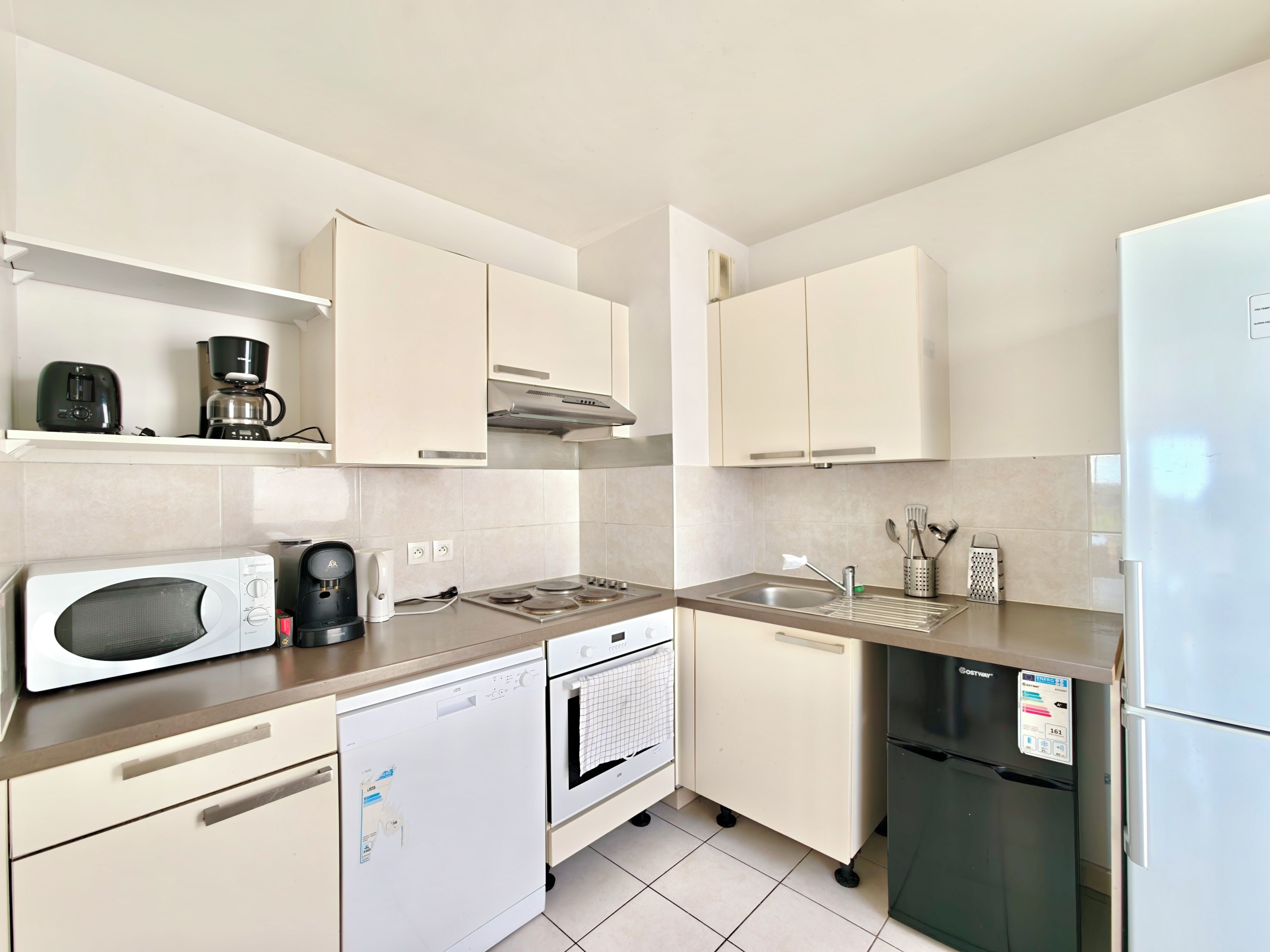 A modern kitchen with white cabinetry, a built-in microwave, a coffee maker, a dishwasher, an oven, a sink, and a refrigerator.