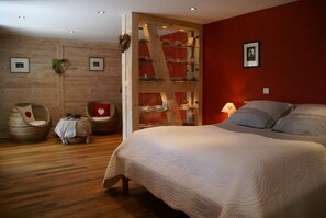 Un dormitorio con una cama grande, un armario de madera, una silla con un cojín con forma de corazón y una mesita con un adorno con forma de corazón.