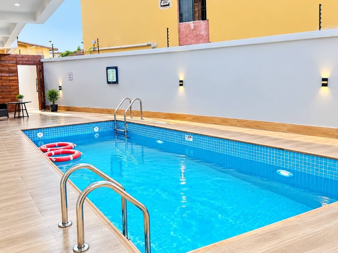 An indoor swimming pool with a clear water surface, a wooden deck surrounding it, and a wall with a light switch and a small plant.
