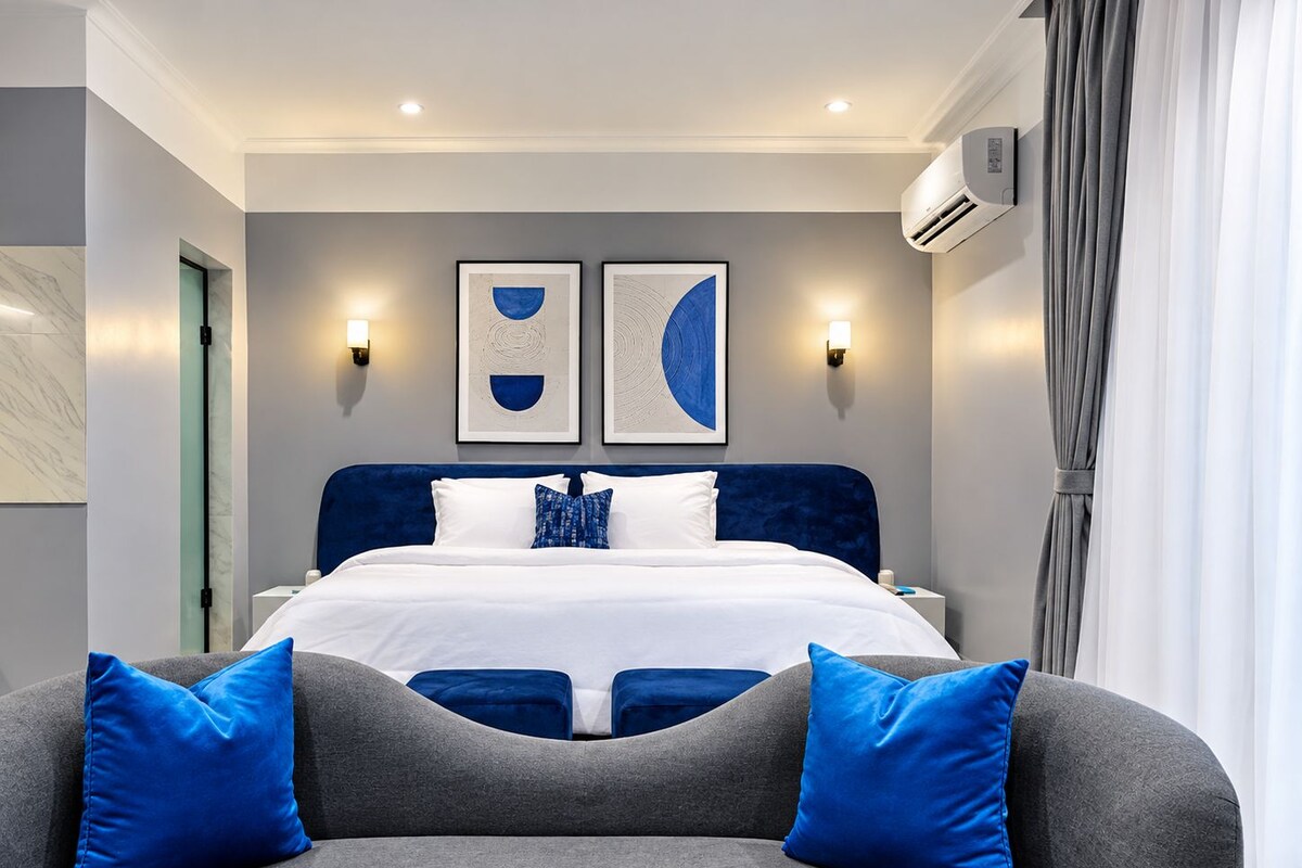 Habitación de hotel moderna con una cama grande, dos obras de arte colgadas en la pared, un sofá gris con cojines azules y un equipo de aire acondicionado.