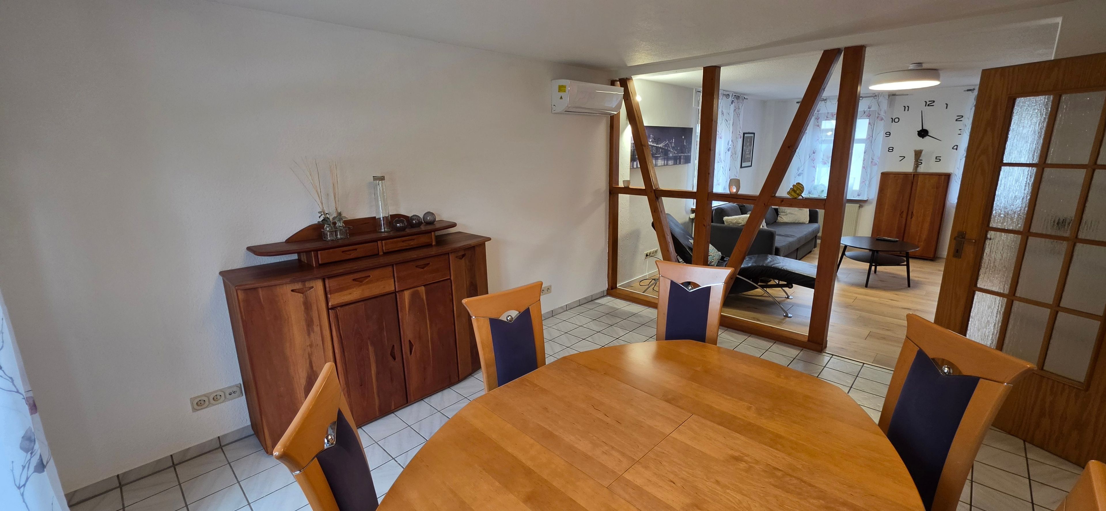 Ein Essbereich mit Holztisch und -stühlen, einem Holzschrank und Blick durch eine Glastür in einen weiteren Raum mit Sofa und Sessel.