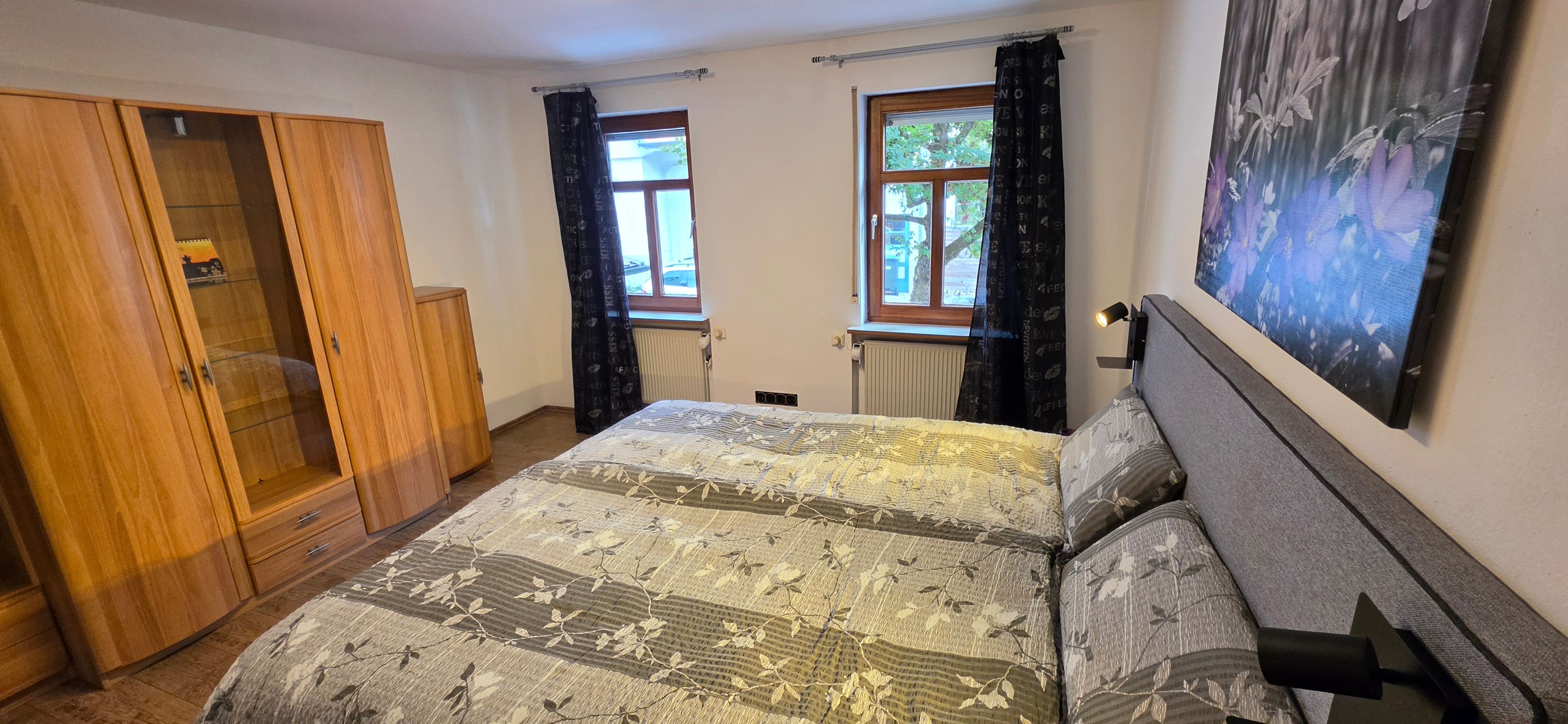 Ein Schlafzimmer mit einem Bett, einem Kleiderschrank, zwei Fenstern mit Vorhängen, einem Heizkörper und einer Wandlampe.