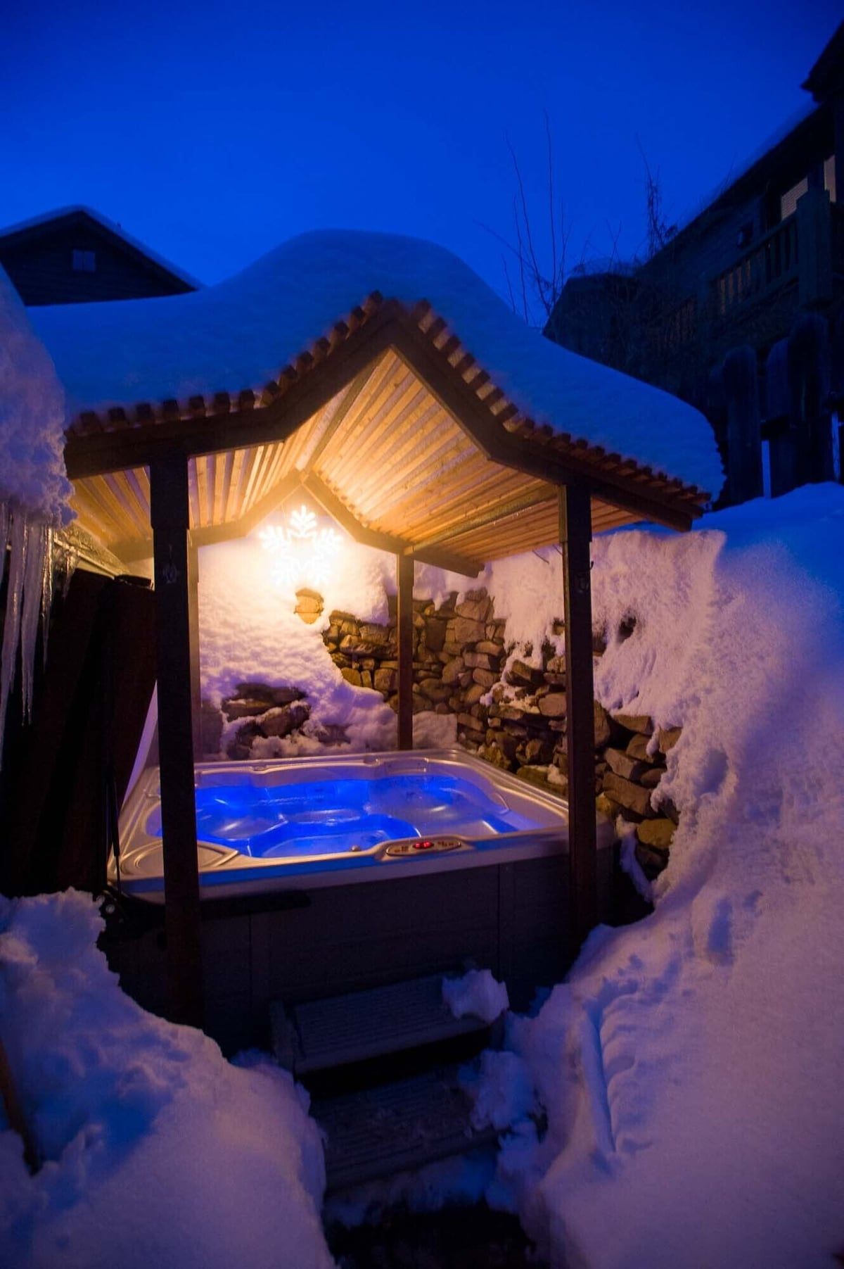 A hot tub surrounded by snow, with a stone wall and a wooden pergola structure.