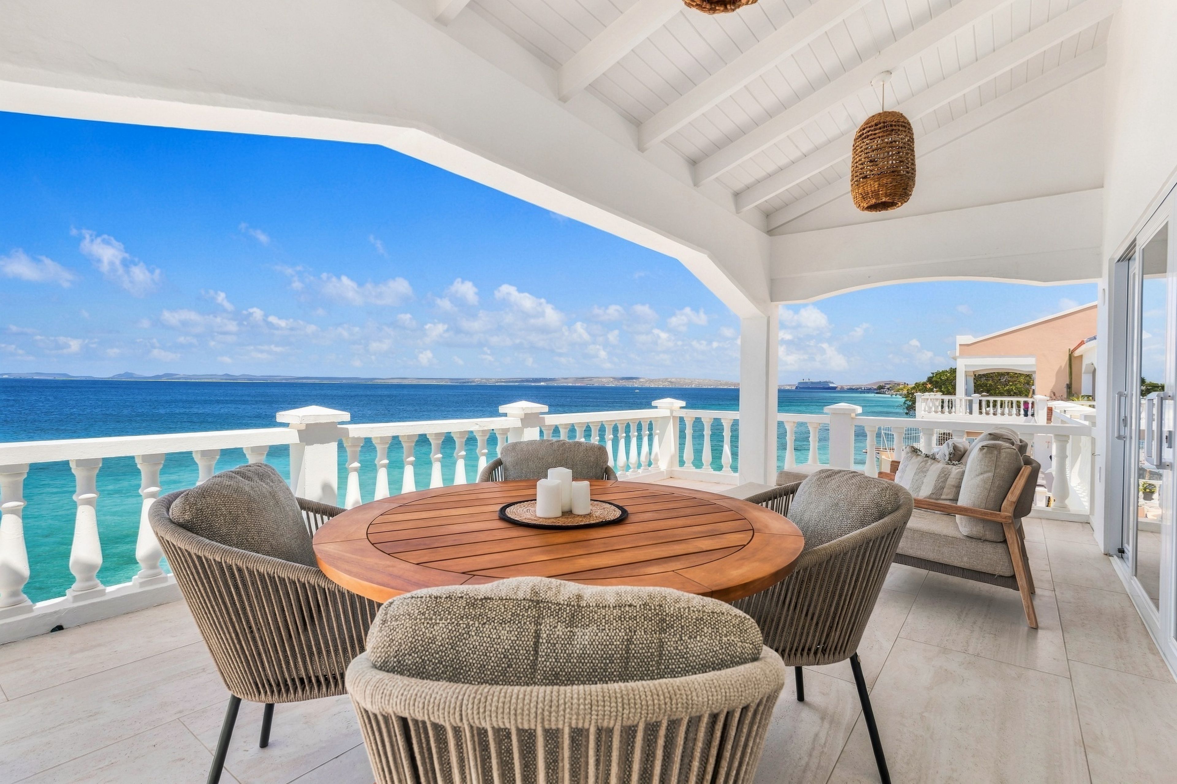 A covered outdoor seating area with a view of the sea, featuring a round wooden table and wicker chairs.