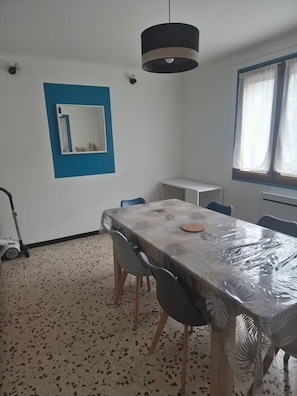 Un coin repas avec une table recouverte d’une nappe à motifs de feuilles, entourée de chaises, d’un luminaire suspendu, d’une petite armoire blanche et d’une fenêtre ornée de rideaux légers.