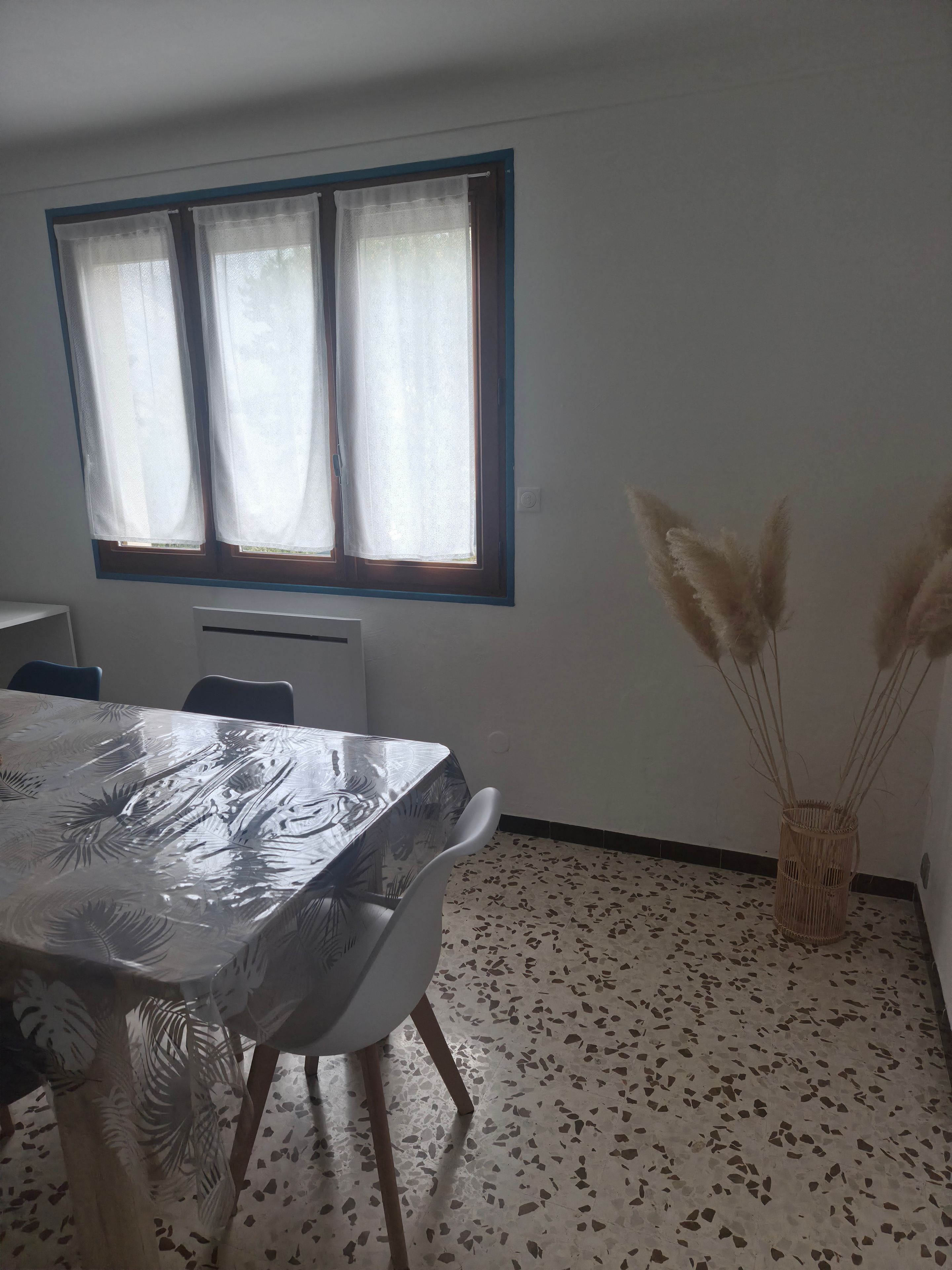 A dining area with a table covered in a clear plastic tablecloth, chairs, a window with curtains, and a vase with pampas grass.
