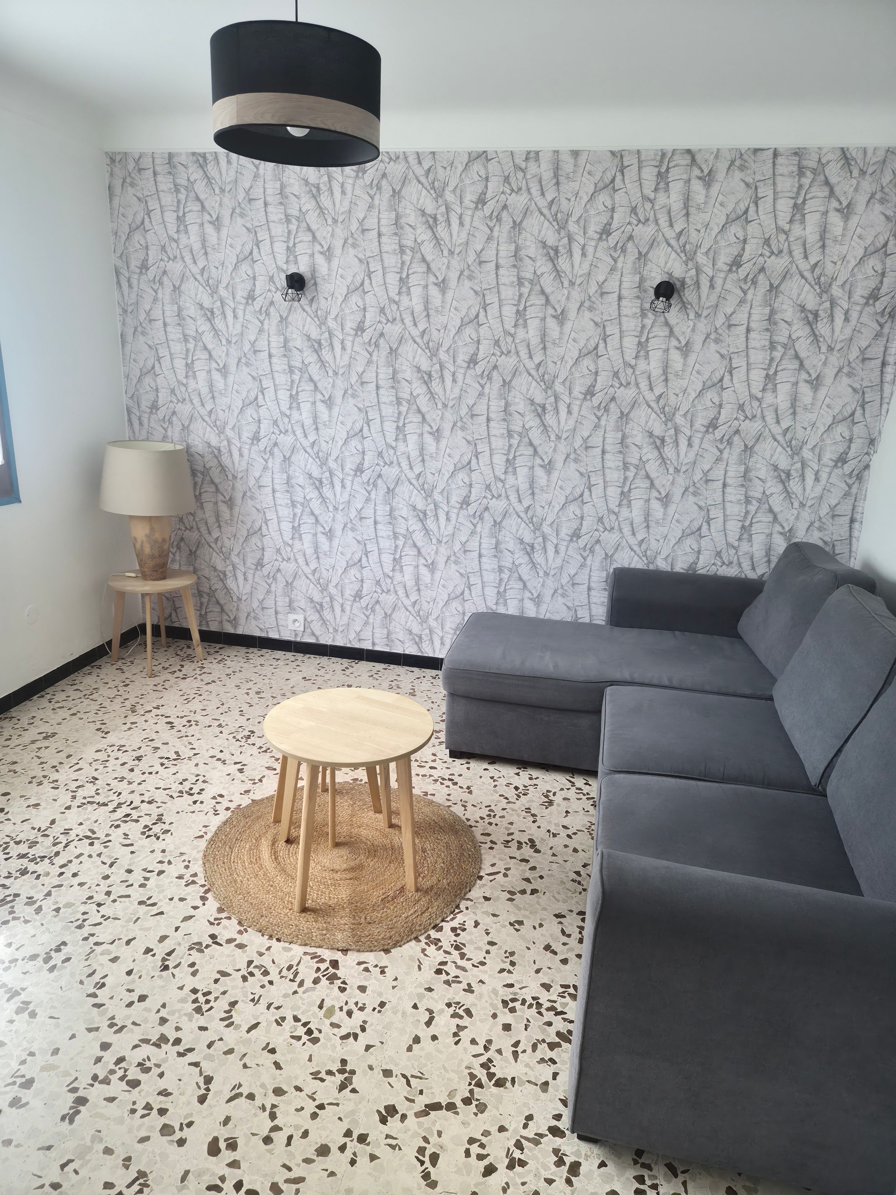 Un salon moderne avec un canapé d’angle gris, une table basse ronde en bois, une lampe et un papier peint à motifs.