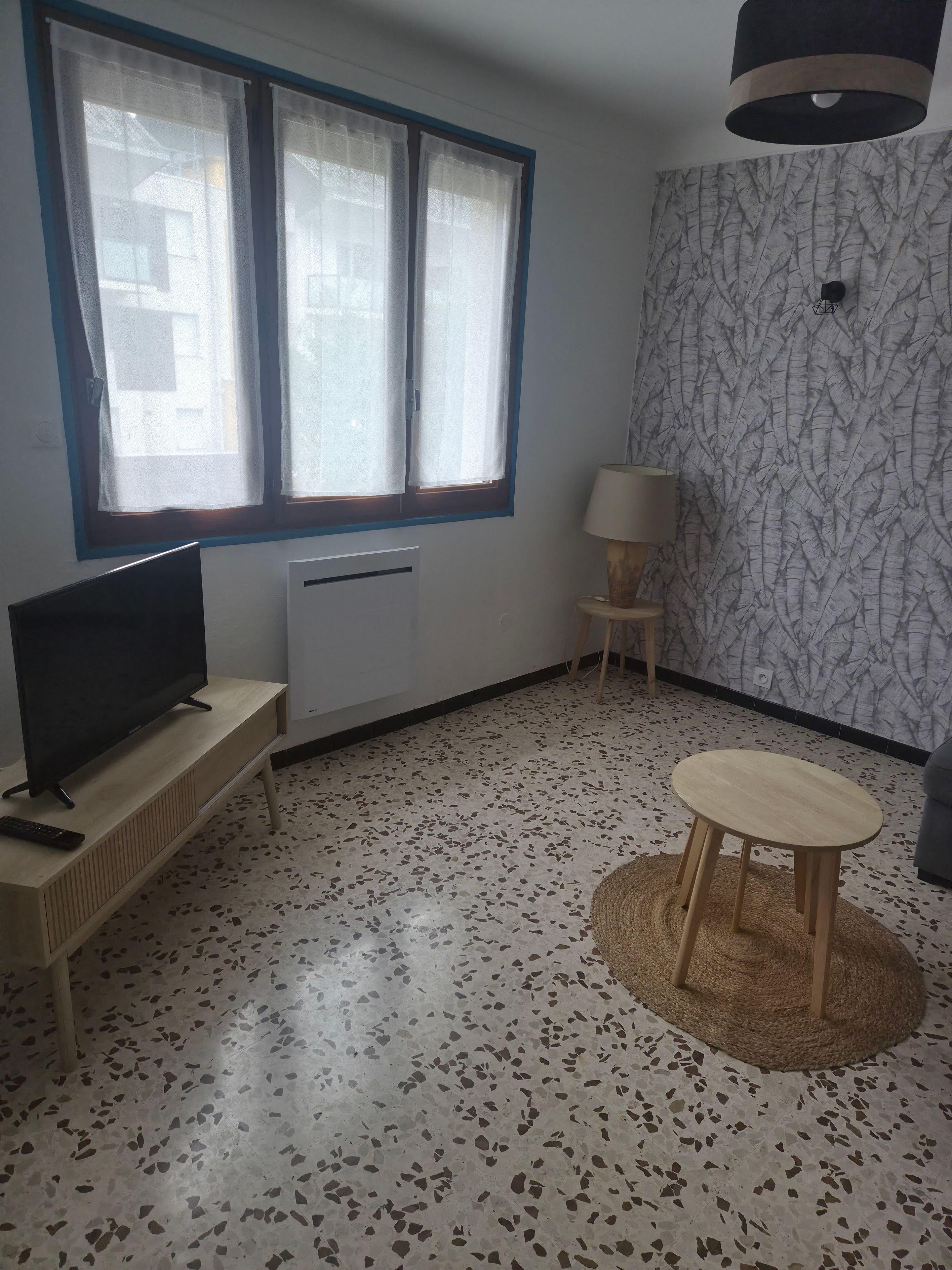 Une pièce avec un téléviseur posé sur un meuble en bois, un radiateur, un mur à motifs, une table ronde et une lampe.
