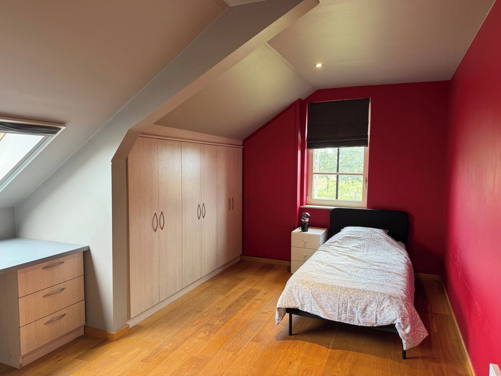 A bedroom with a sloped ceiling, a bed, a nightstand, a wardrobe, and a window with a view.