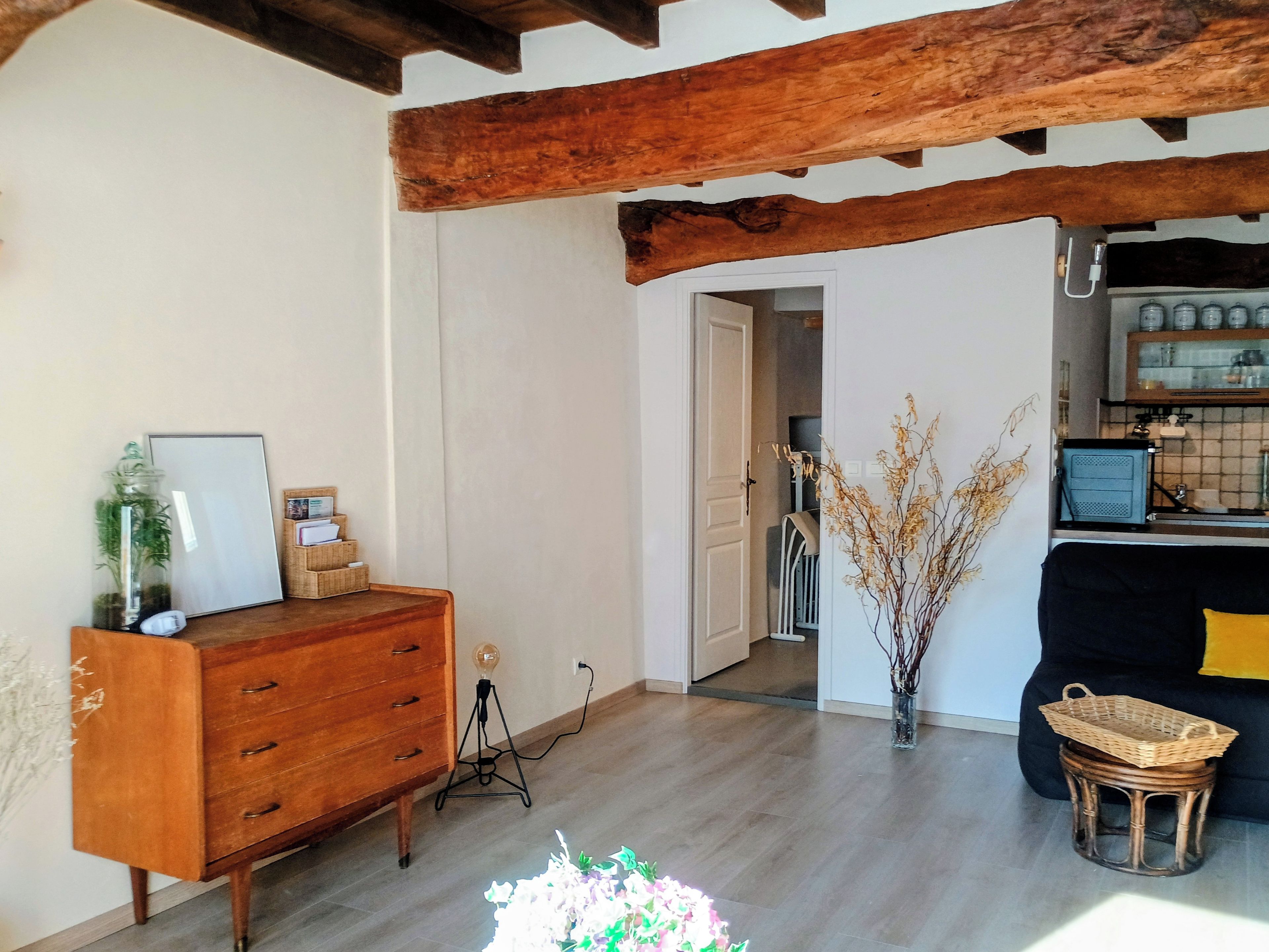 Une pièce comprenant une commode en bois, une lampe, un vase contenant des branches séchées et un panier en osier posé sur un tabouret.