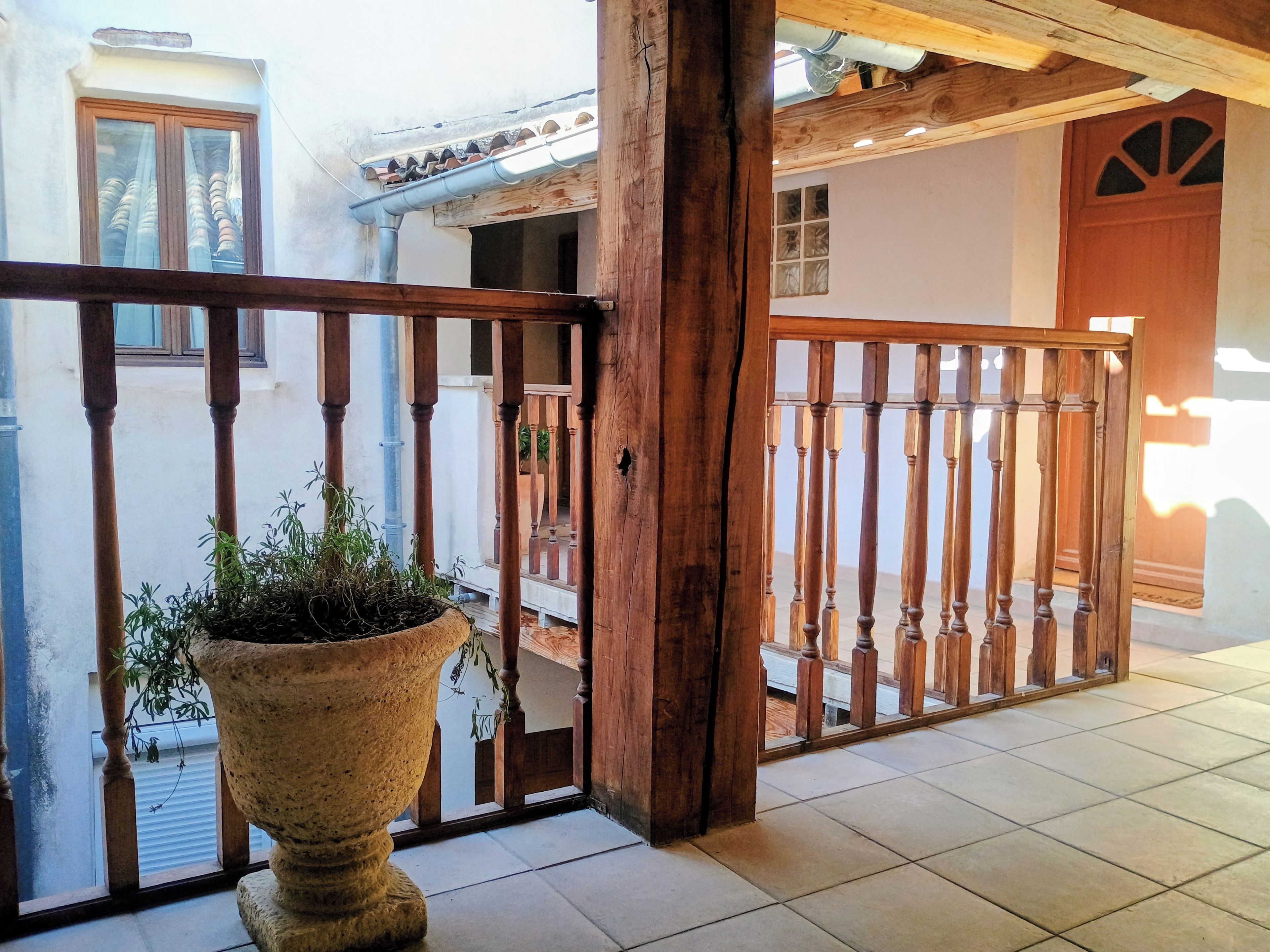 Un balcon en bois orné d’un pot de fleurs, un sol carrelé et une vue sur un immeuble avec une fenêtre et une porte.