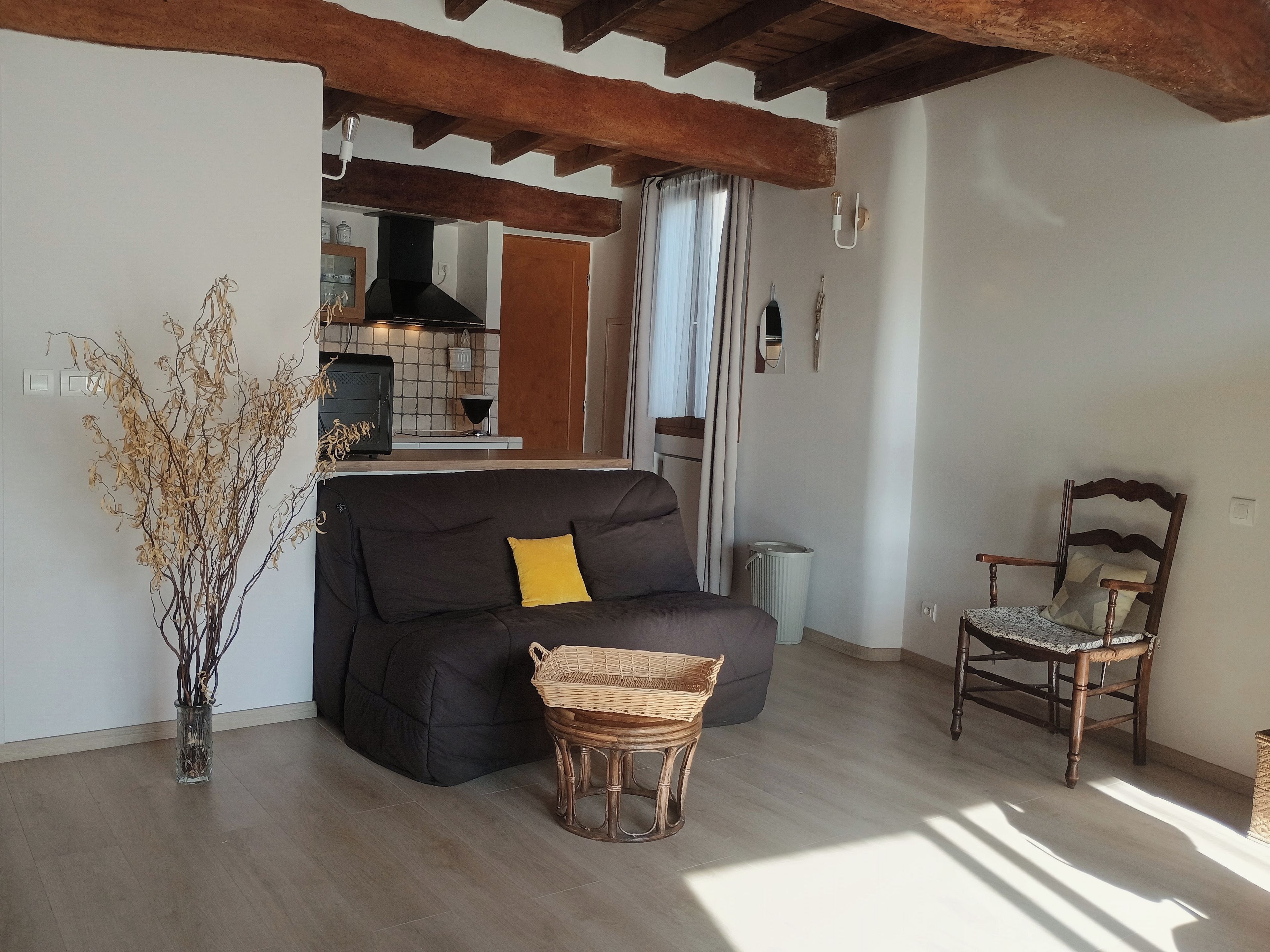 Un salon avec un canapé foncé, une table d’appoint en osier, une chaise en bois et un vase contenant des branches séchées.