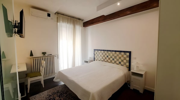 A hotel room with a bed, a desk, a chair, a radiator, and a window with curtains. - Casa Alba - Mara Room (Caltagirone)