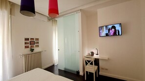 A hotel room with a bed, a desk with a chair, a radiator, a television mounted on the wall, and a door leading to another room. - Casa Alba - Camera Elisa (Caltagirone)