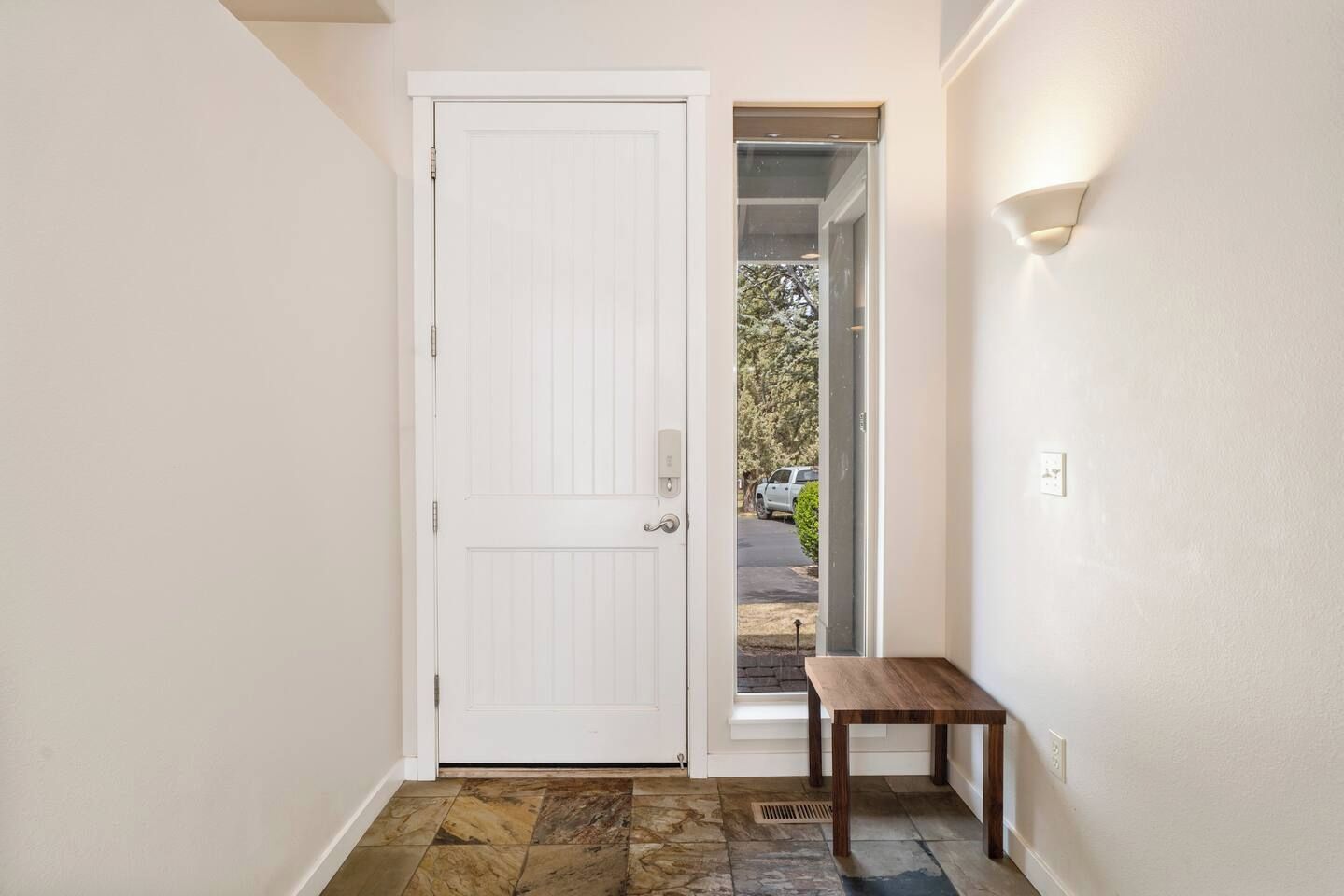 Un couloir avec une porte blanche, un petit banc en bois et une vue sur une allée où est garé un véhicule.