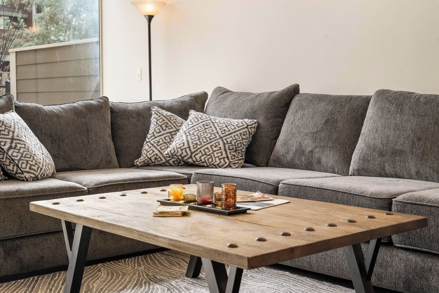 Un canapé d’angle gris avec des coussins à motifs, une table basse en bois sur laquelle sont posées des boissons et des magazines, et une lampe sur pied en arrière-plan.
