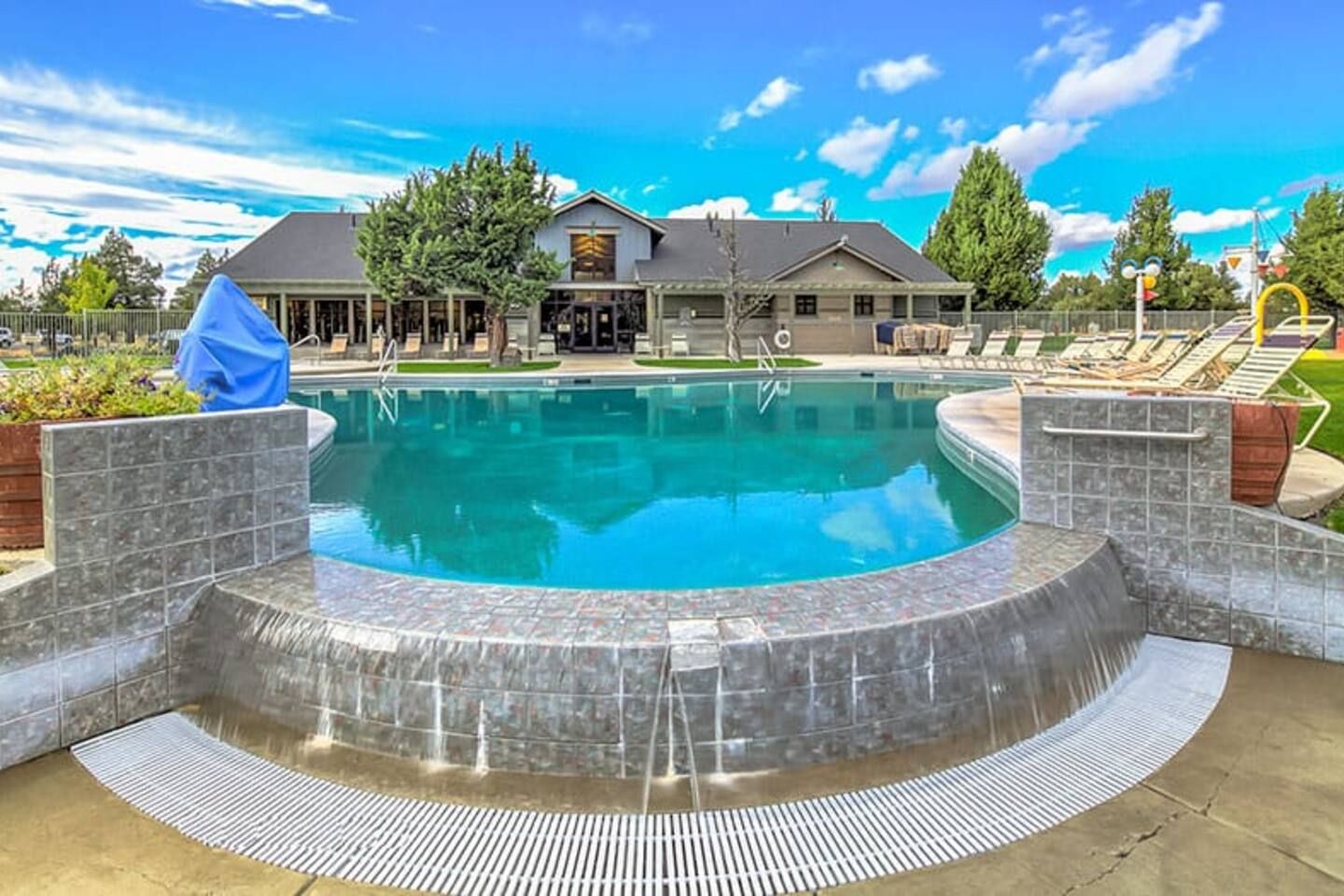 Une piscine bordée de pierres, un espace de détente couvert et un bâtiment en arrière-plan.