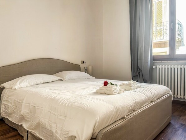 A neatly made bed with white linens, a red rose, and towels, in a room with a radiator and a window with curtains.