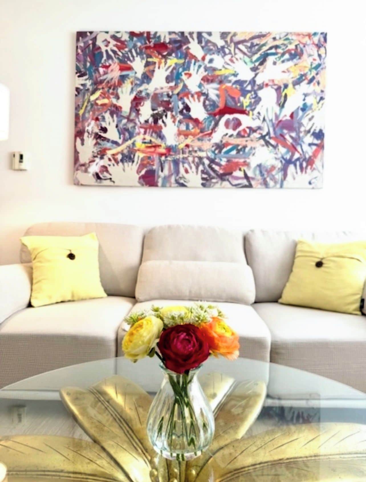 A living room with a sofa, yellow cushions, a glass coffee table with a vase of flowers, and a colorful abstract painting on the wall.