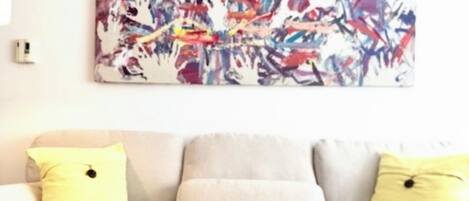 A living room with a sofa, yellow cushions, a glass coffee table with a vase of flowers, and a colorful abstract painting on the wall.