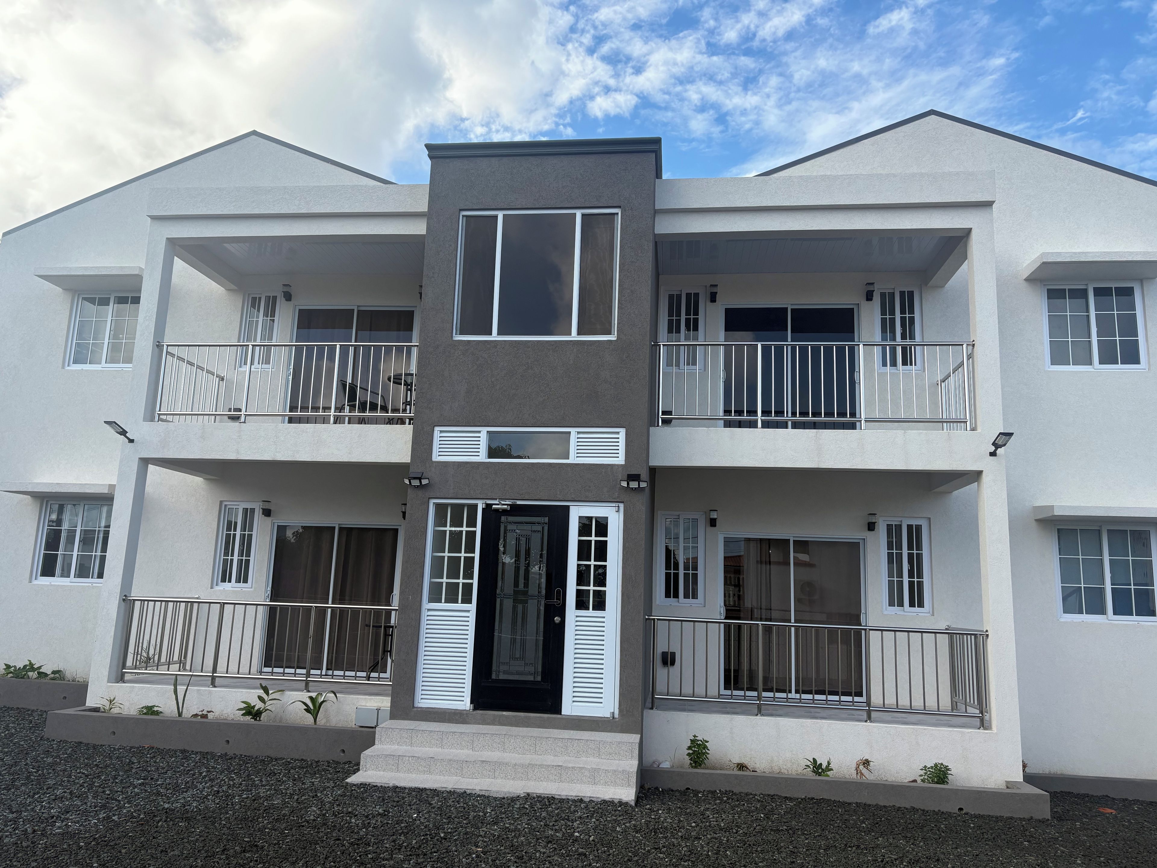 A multi-story residential building with balconies, a central entrance, and a gravel driveway.