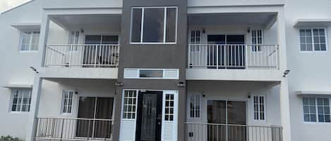 A multi-story residential building with balconies, a central entrance, and a gravel driveway.