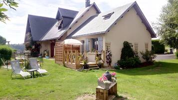 A two-story house with a sloped roof, a wooden deck, and a garden with chairs and potted plants.