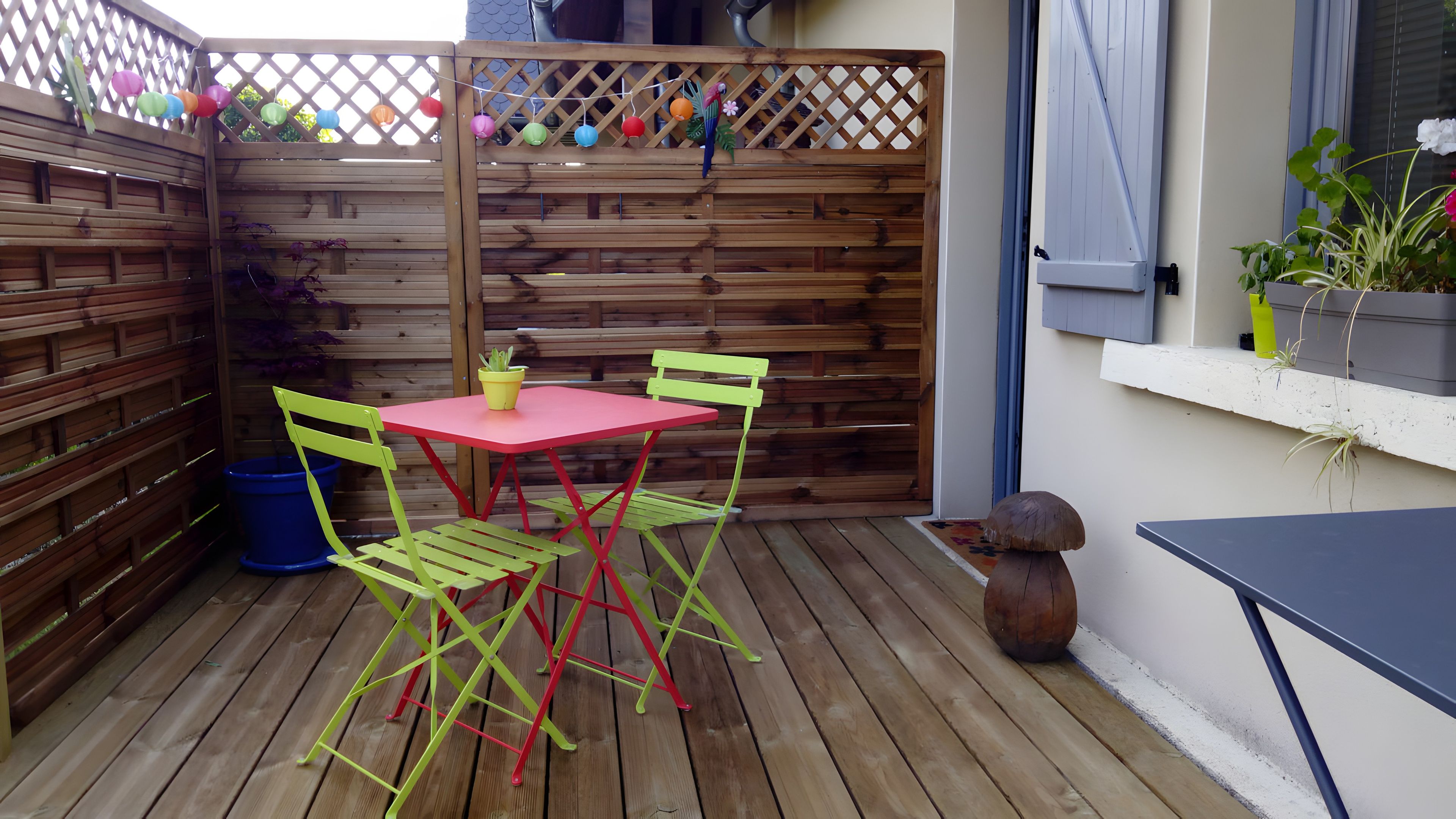 Eine Holzterrasse mit einem roten Tisch und grünen Stühlen, einem blauen Mülleimer und einem Holzzaun mit bunten hängenden Dekorationen.