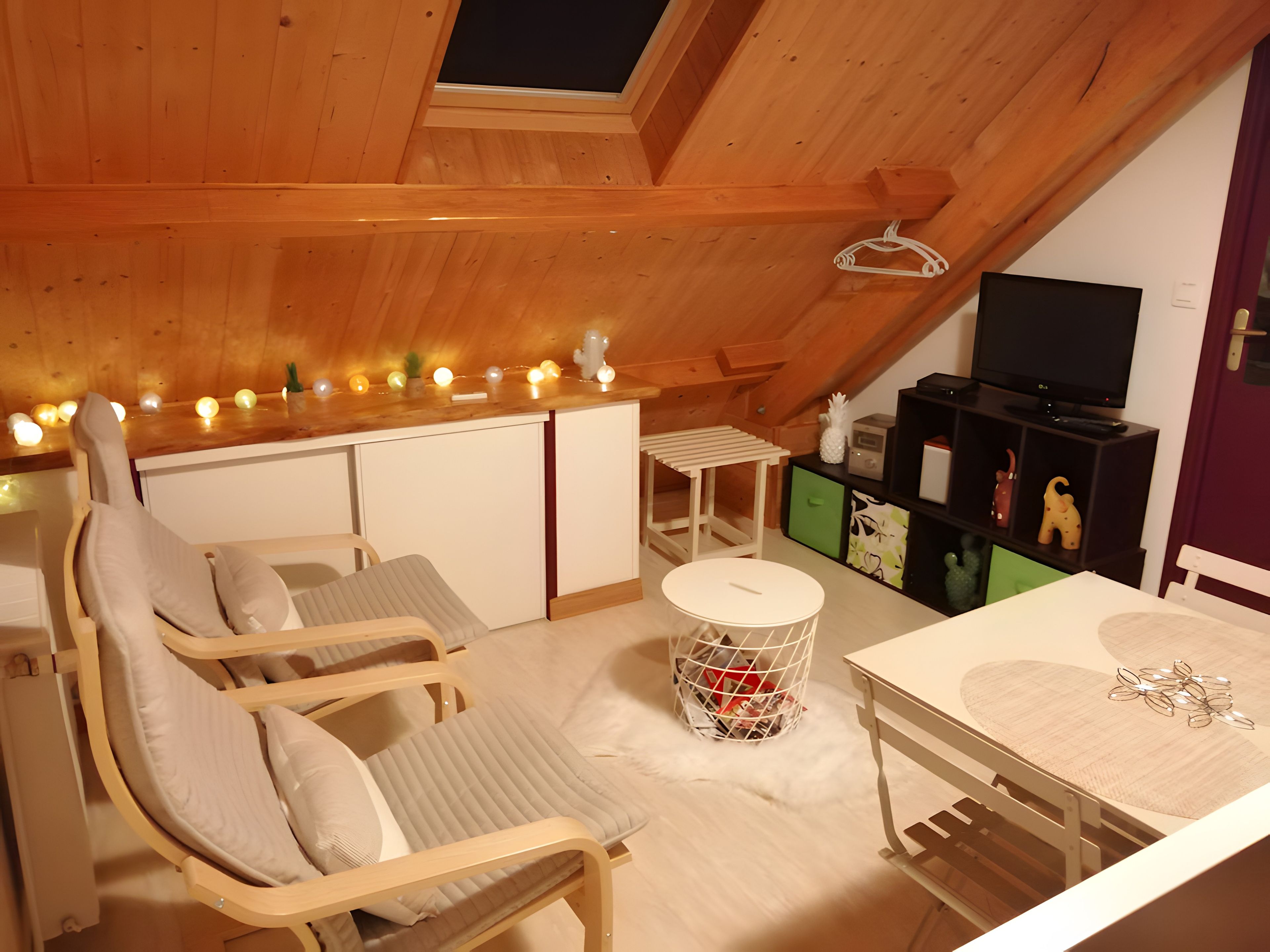 Een gezellig zolderkamer met schuine wanden, houten balken, een televisie, een eettafel en een zithoek met stoelen.