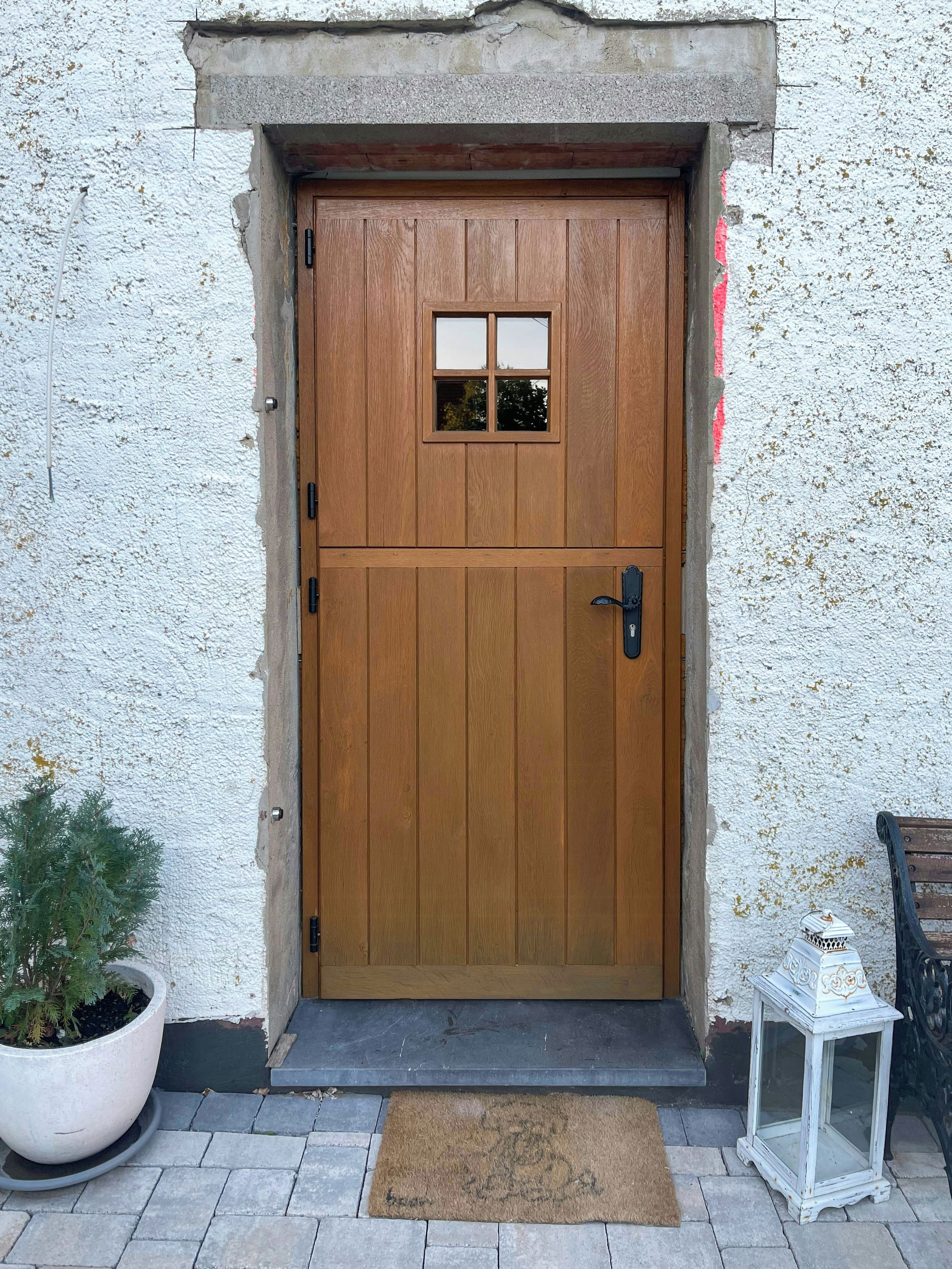 Eine Holztür mit kleinem Fenster, einer Fußmatte und einem Blumentopf daneben.