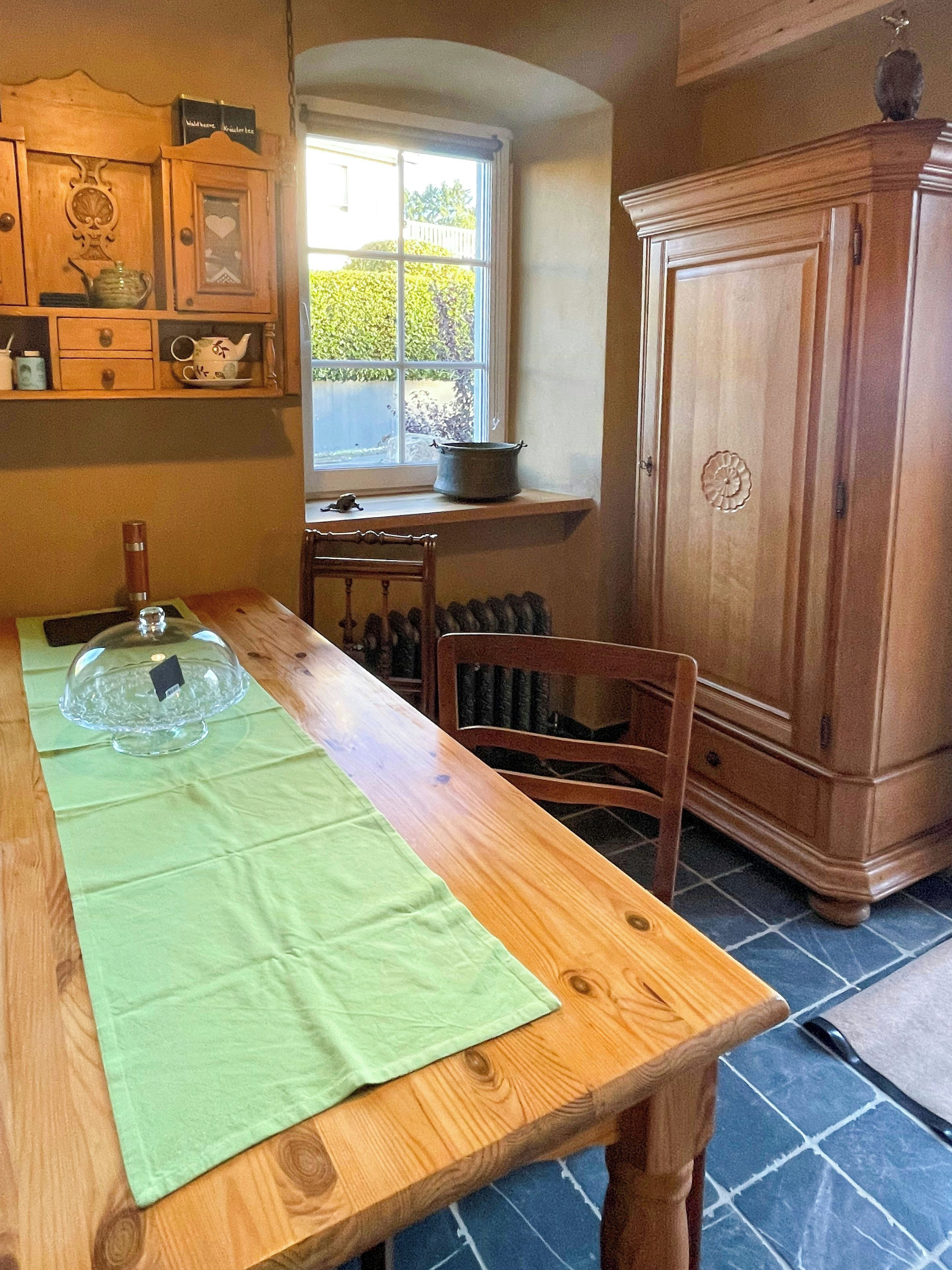 Een houten eettafel met een groene tafelkleed, een transparante glazen taartetafel en een raam met uitzicht op groen.