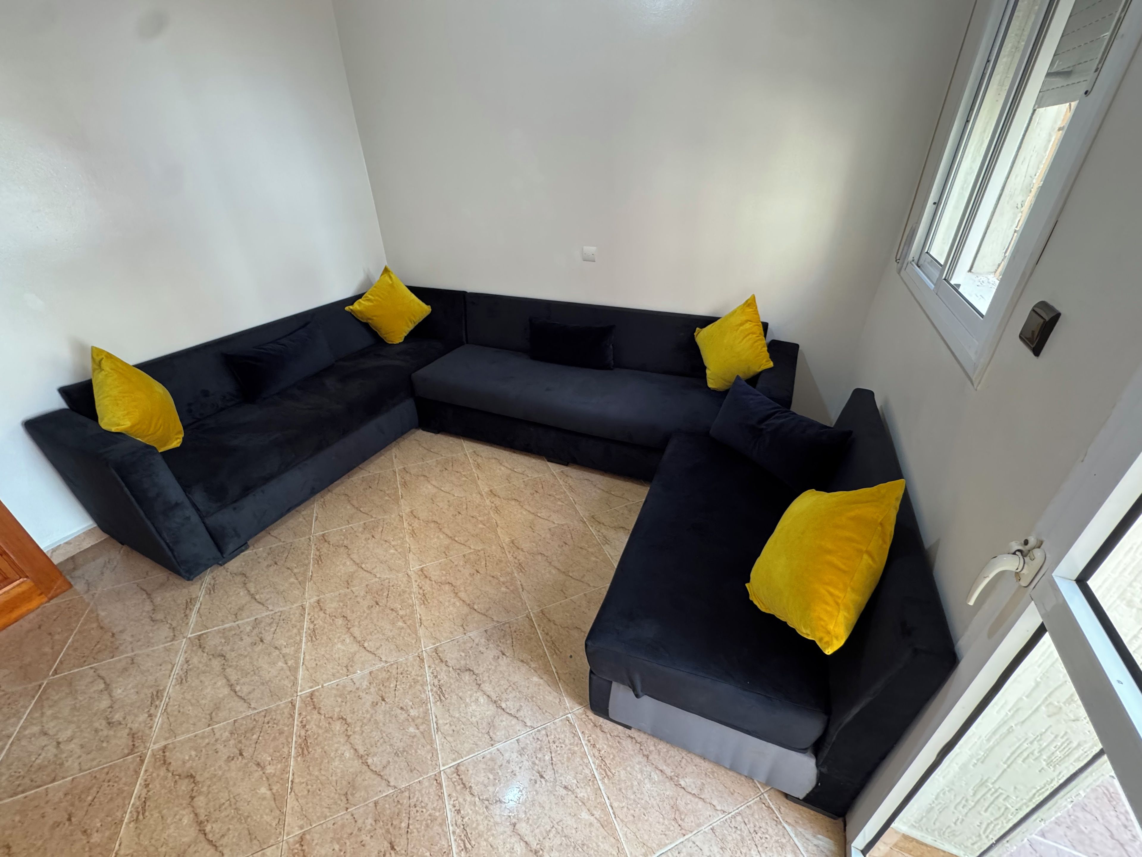 Una sala de estar en esquina con un sofá seccional negro, almohadas amarillas y piso de baldosas.