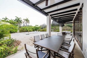 Patio esterno con un tavolo lungo e sedie, immerso nel verde e palme.