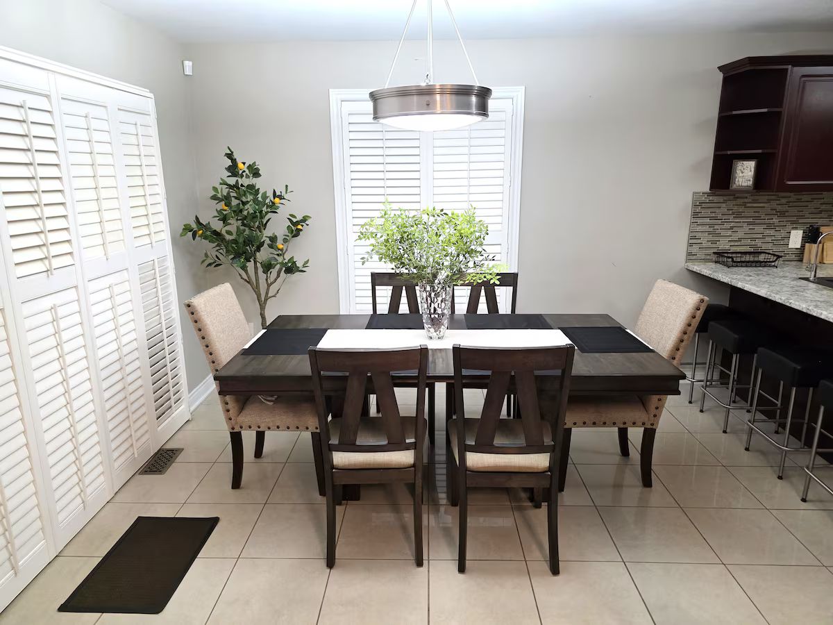 A dining area with a rectangular table, six chairs, a potted plant, a hanging light fixture, and a kitchen counter with bar stools.