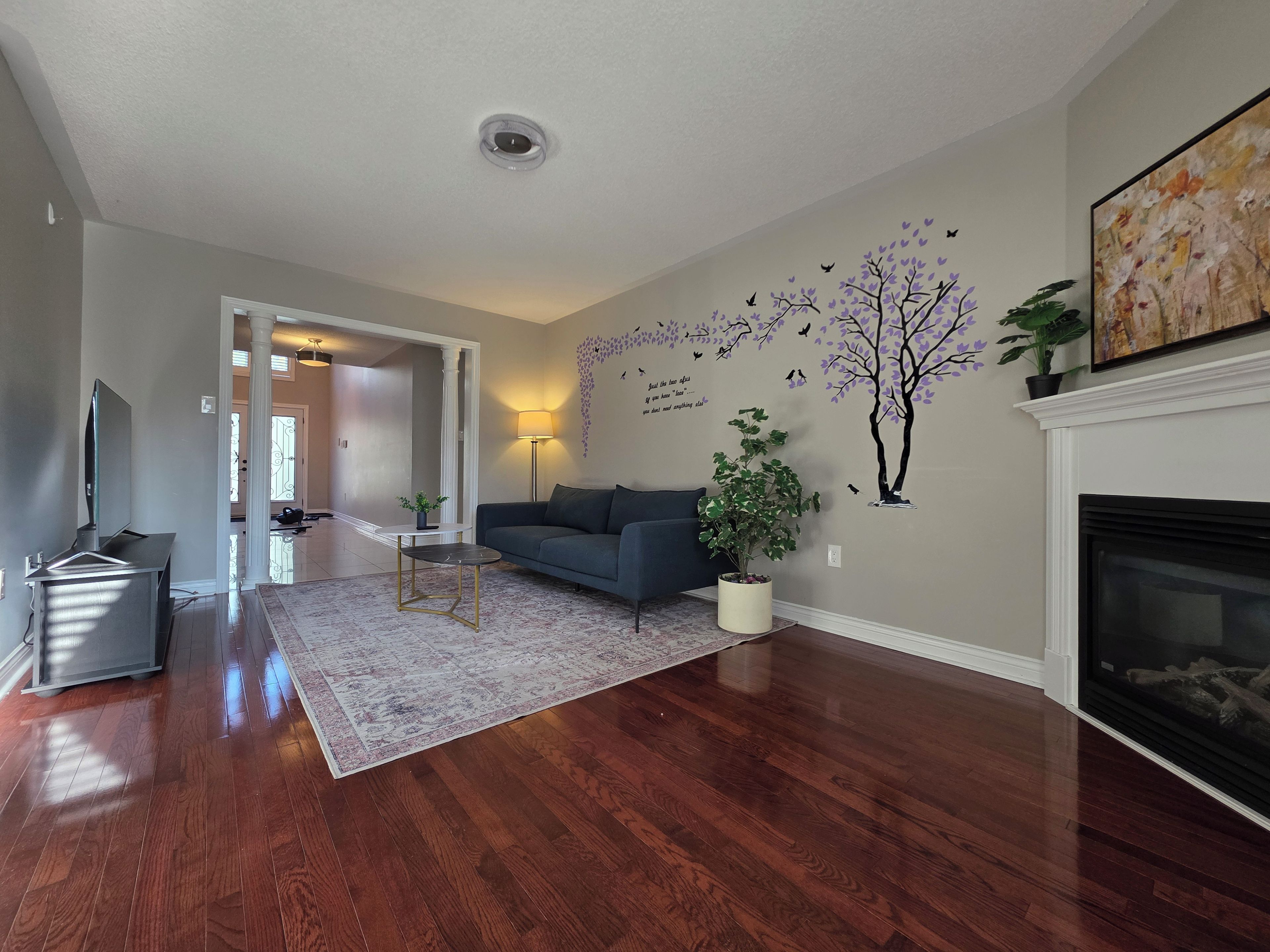A living room with hardwood flooring, a sofa, a coffee table, a TV, a fireplace, and a wall with a tree decal.