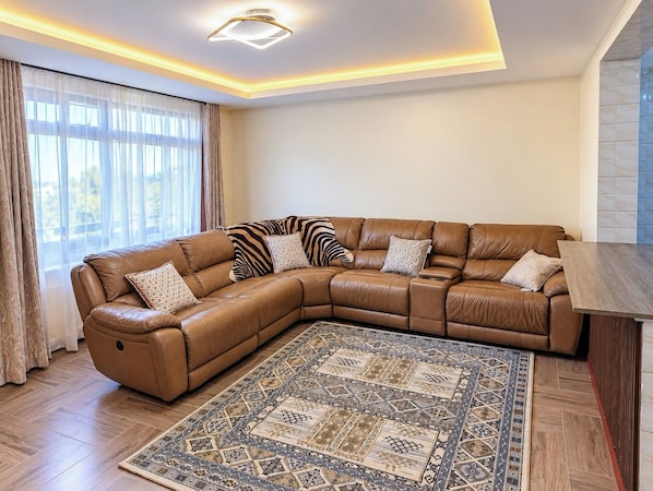 A L-shaped leather sofa with multiple cushions, a patterned rug, and a wooden side table in a well-lit room with a large window.