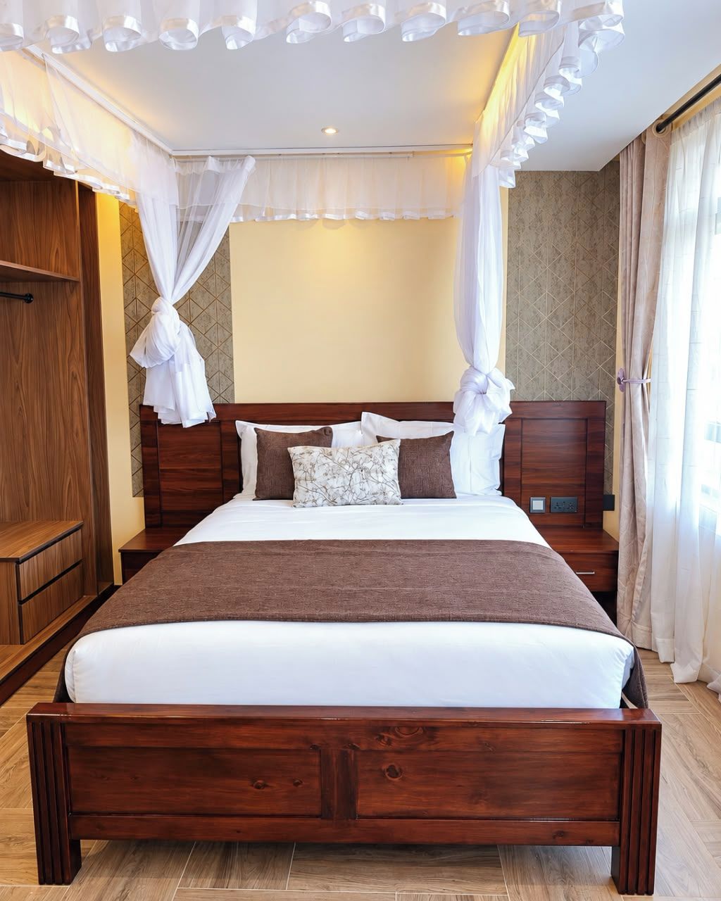 A large bed with a wooden frame, white bedding, and multiple pillows. There is a wooden nightstand beside the bed, and a wooden wardrobe to the left.