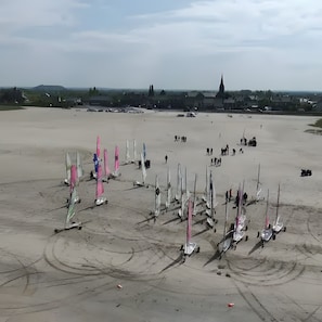 Une vaste étendue de sable où sont amarrés de nombreux voiliers, une église visible au loin et un ciel dégagé.