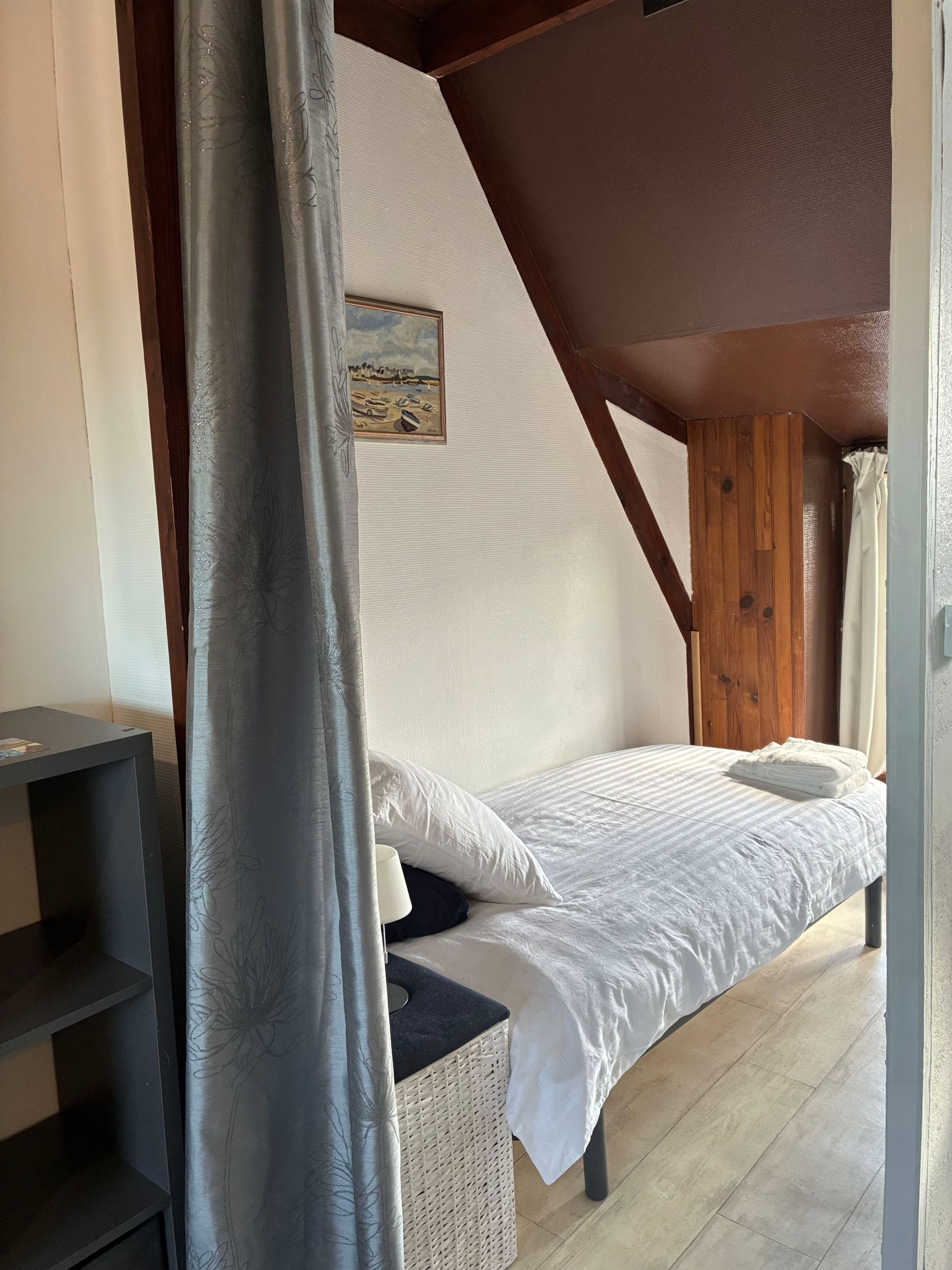 Ein Einzelbett in einem Zimmer mit schrägem Dach, einem kleinen Tisch und einem Korbstuhl.