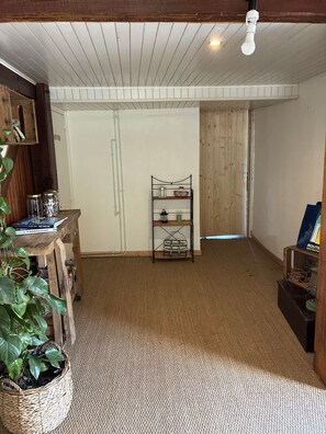 Una habitación con suelo de madera, techo blanco, estantería con libros y frascos, una planta y una puerta que da a otra habitación.