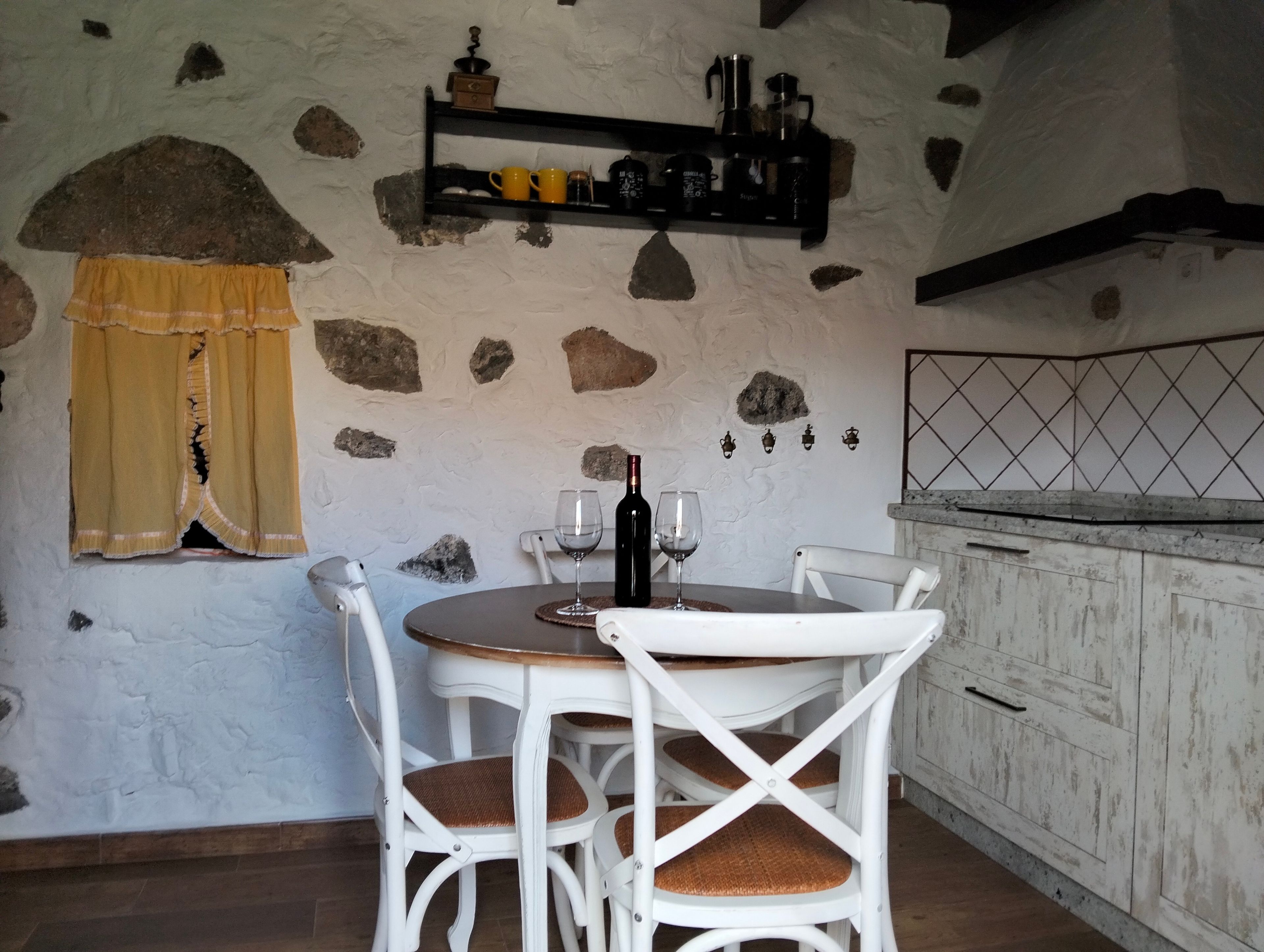 Een landelijke keuken met een stenen muur, een houten tafel met stoelen en een plank met keukengerei.