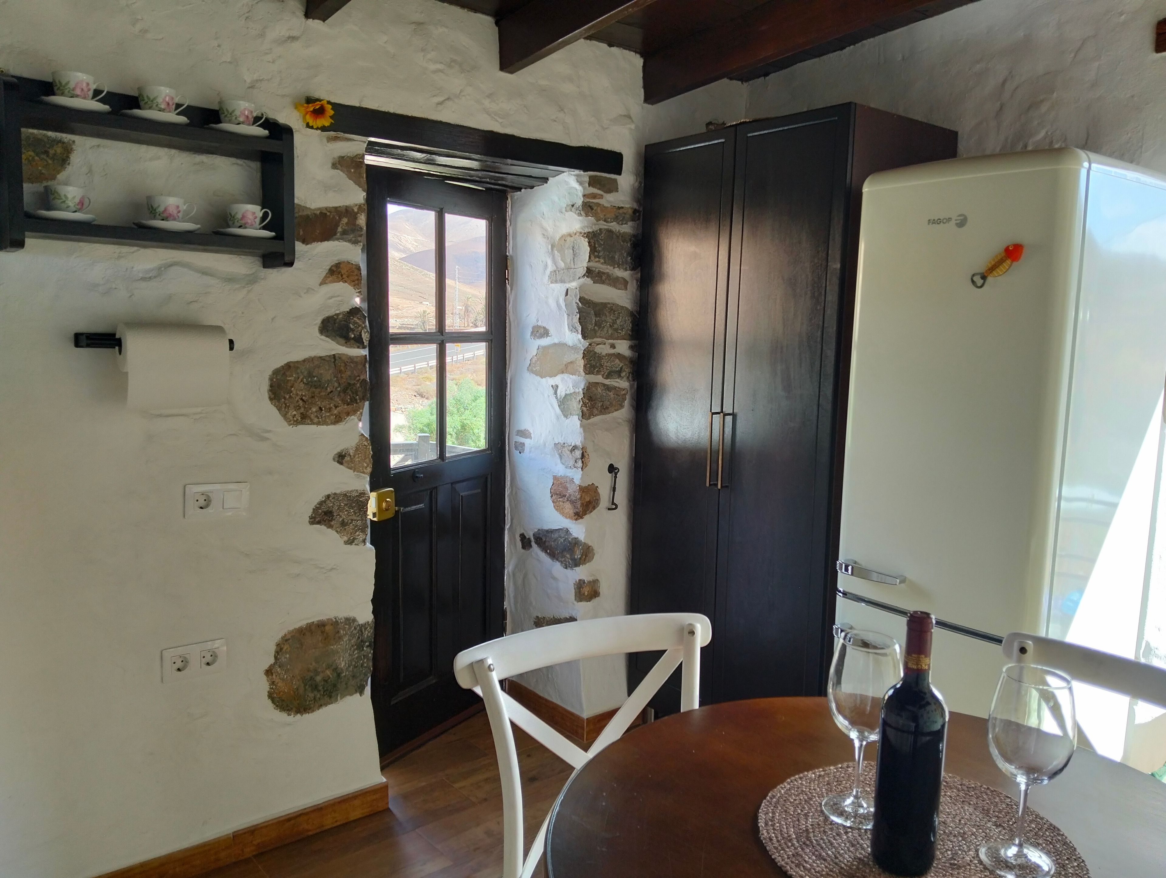 Eine Küche mit einer Steinwand, einer schwarzen Tür, einem weißen Stuhl, einem Holztisch, einem weißen Kühlschrank und einer Weinflasche.