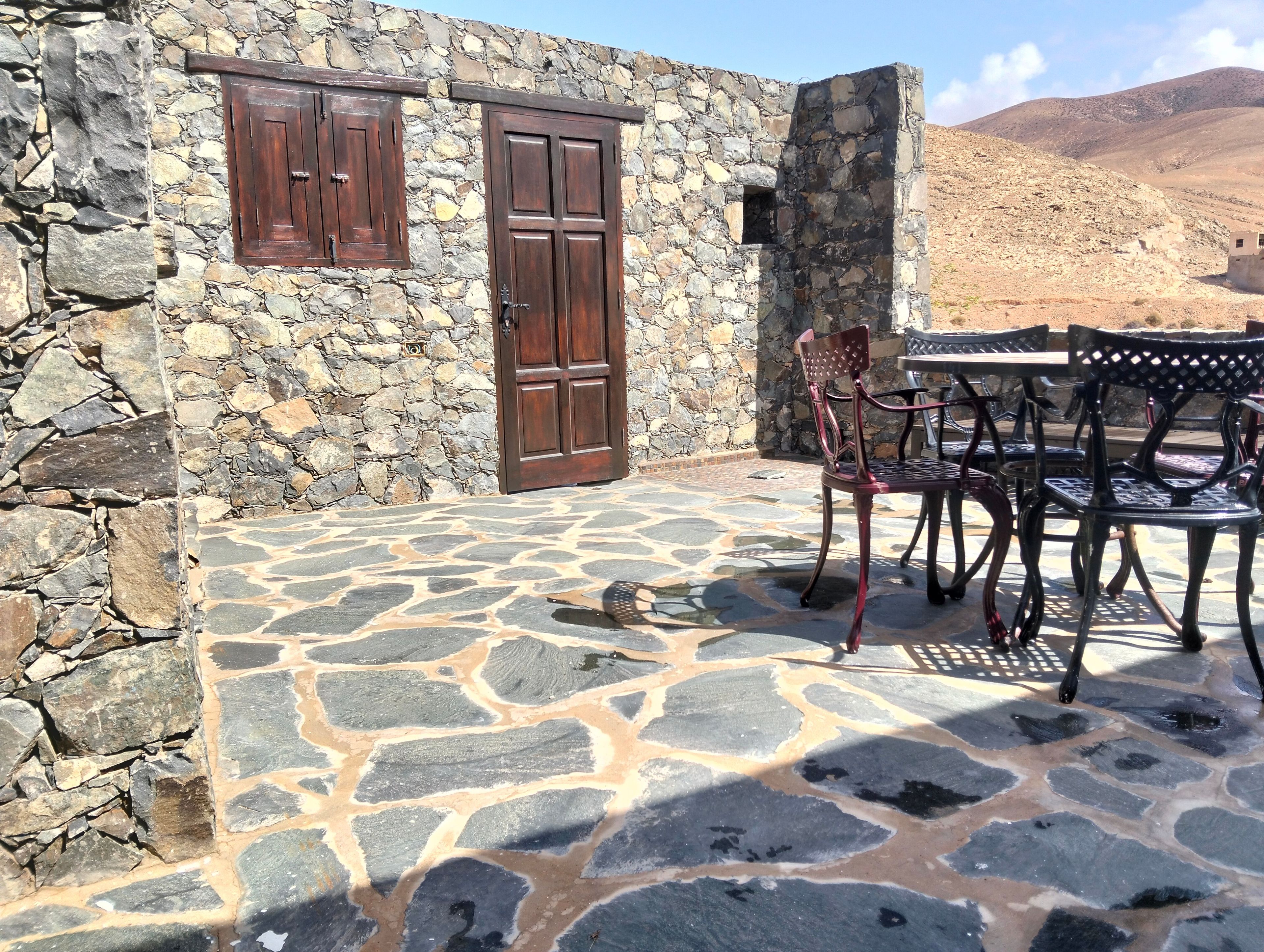 Un bâtiment en pierre doté de deux portes en bois et d’une terrasse en pierre avec une table et des chaises.